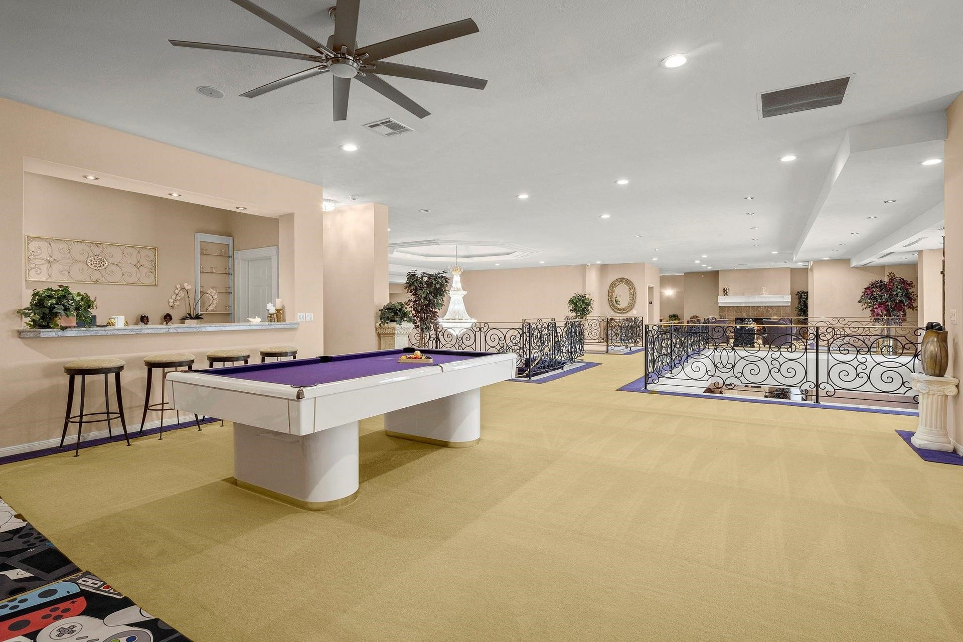 You're looking at a spacious entertainment room featuring a pool table, a bar area with stools, and elegant wrought-iron railings. The room has recessed lighting and a large ceiling fan, providing a comfortable and inviting space for leisure and social gatherings.