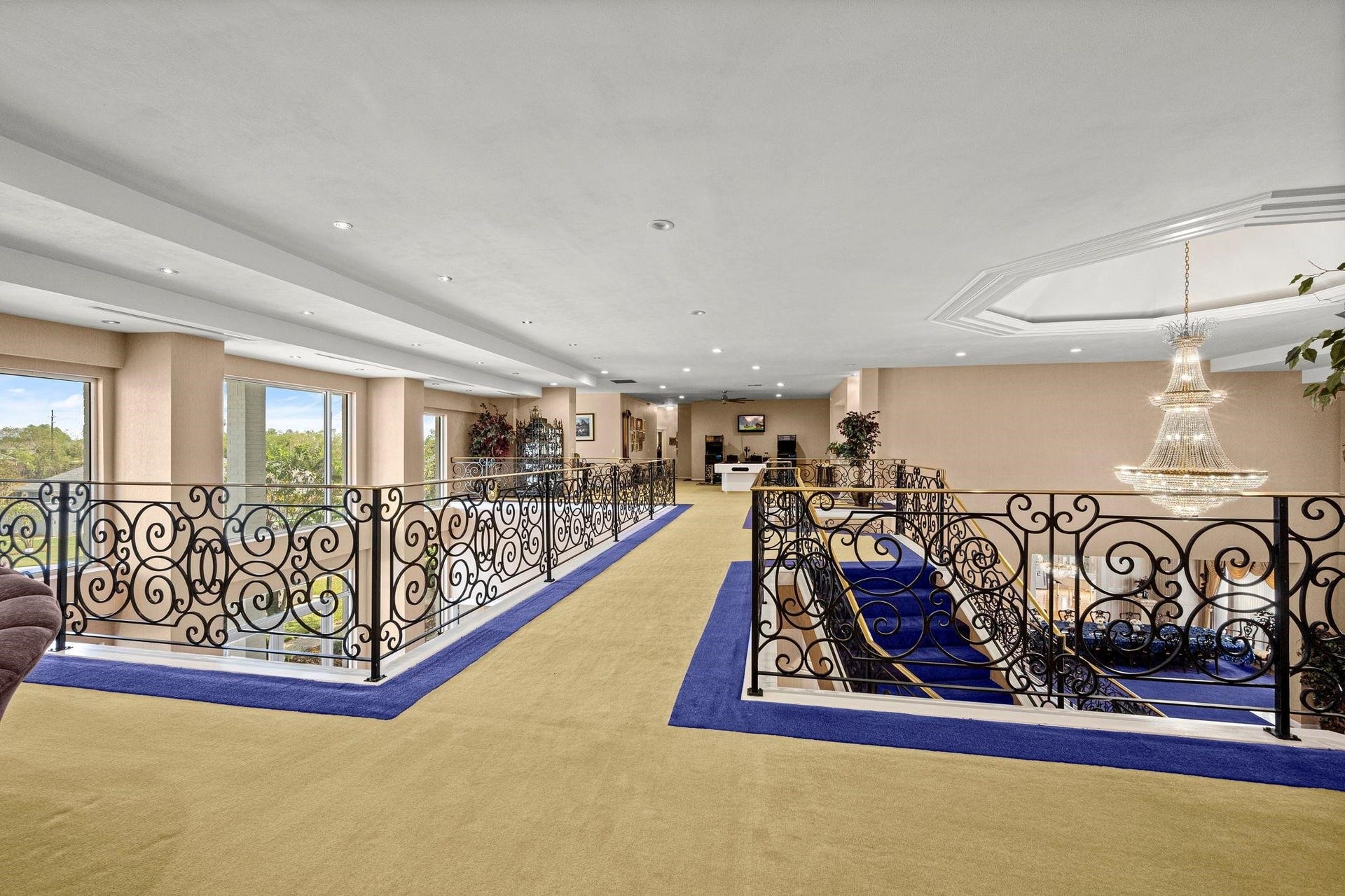 This is a spacious, well-lit interior space featuring an elegant open floor plan with a grand staircase, ornate metal railings, and large windows that provide ample natural light and views of the surrounding area. A classic chandelier adds a touch of luxury to the room.