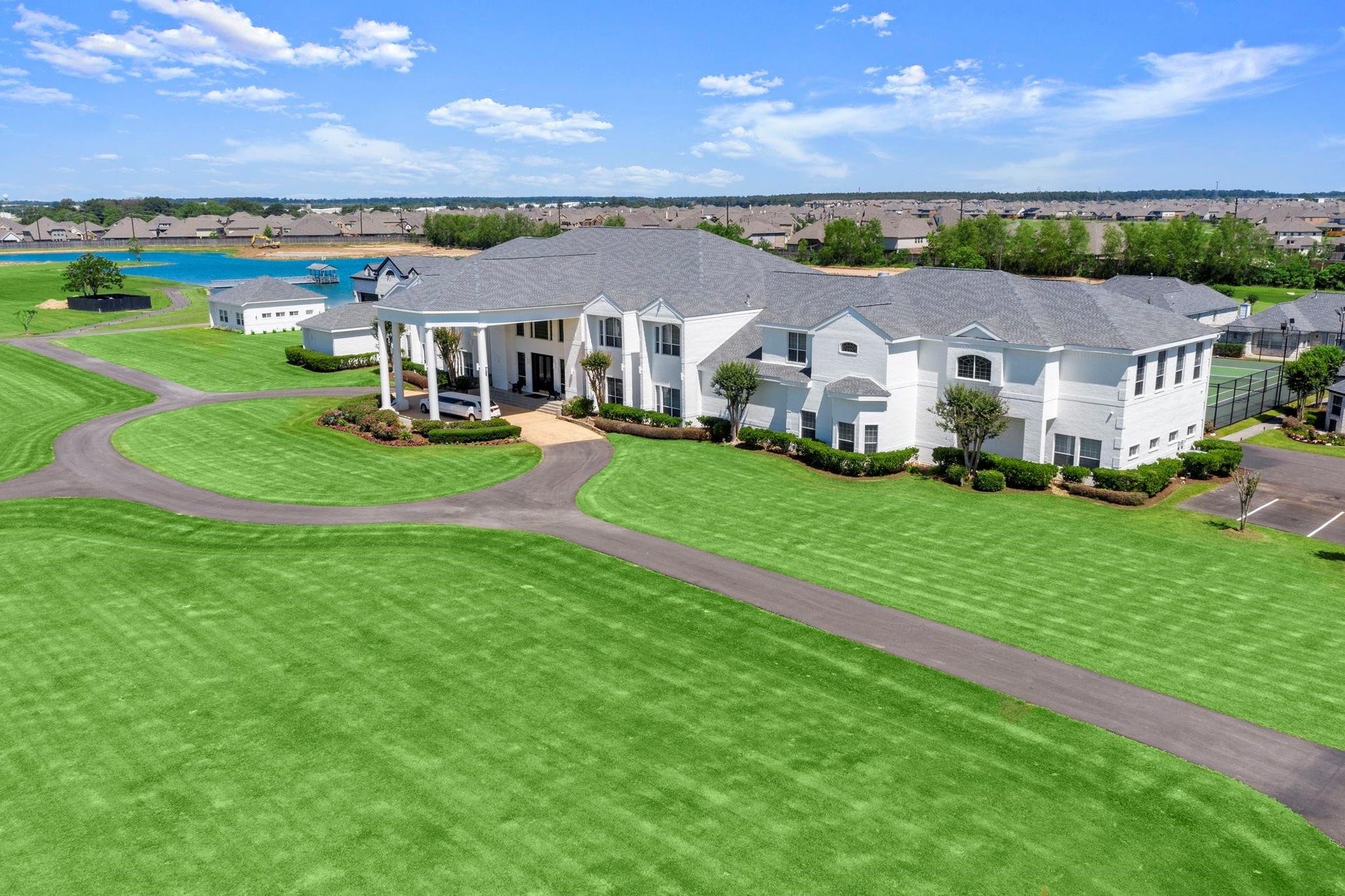 This is an aerial view of a large, multi-story white house with a prominent columned entrance, surrounded by expansive, well-manicured lawns. The property includes a circular driveway, an attached multi-car garage, and is located in a suburban area with nearby water features and other homes in the distance.