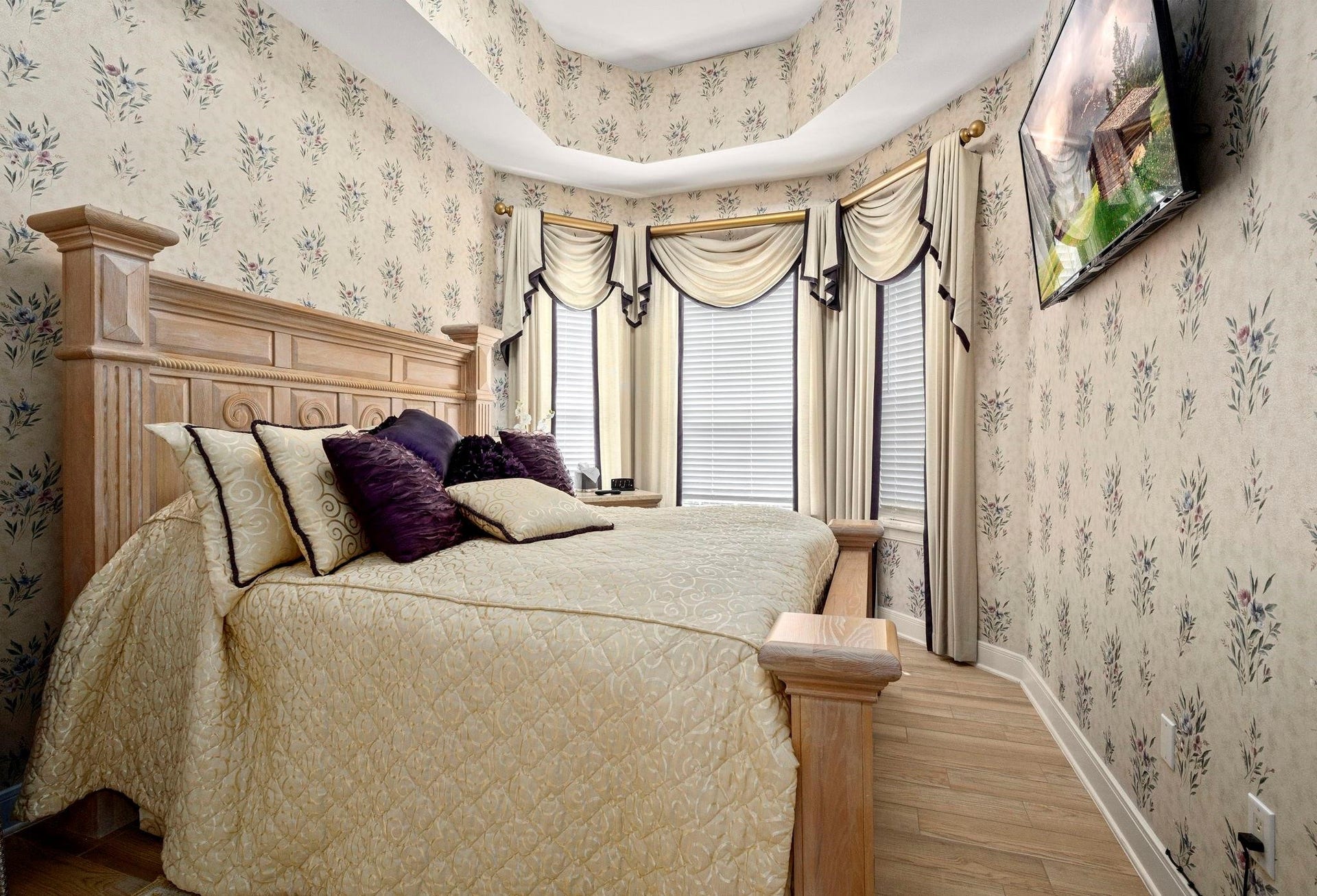 This is a traditional-style bedroom featuring floral wallpaper, a wooden headboard, elegant window draperies, and a matching bedspread with decorative pillows. The room has a classic charm and includes a flat screen T.V. on the wall.