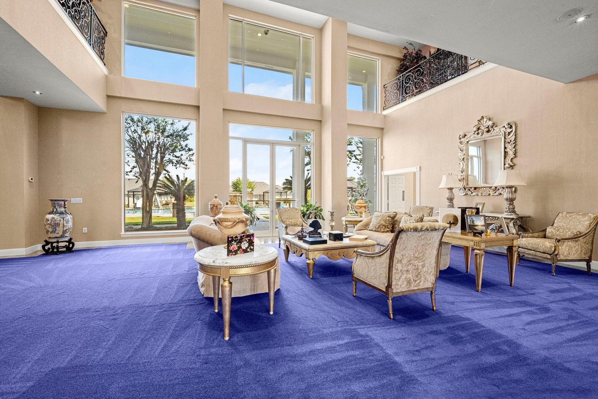This is a spacious living room with high ceilings, large windows providing ample natural light, and a view of the outdoors. It features a bold purple carpet and classic-style furniture, with a second-story indoor balcony overlooking the space.