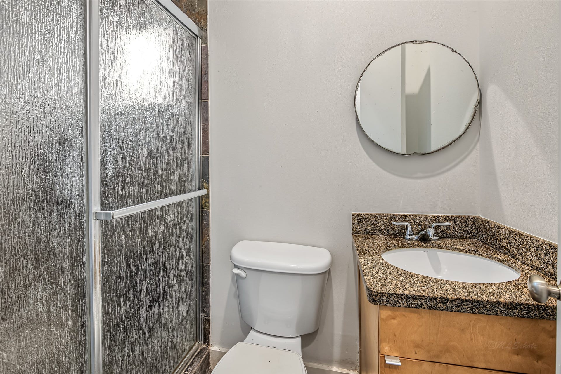 1555 Birdsall Street, #A, Houston, TX 77007 The ground-floor en-suite full bathroom includes a walk-in shower with privacy glass, slate-look tile framing, a granite-topped vanity, and a chic circular mirroroffering complete privacy and convenience for visitors or ground-level living.