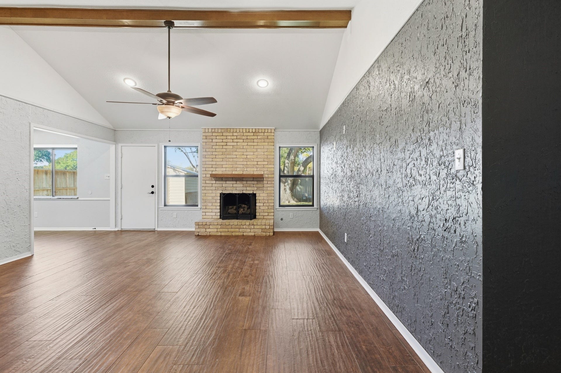3721 Avenue R, Rosenberg, TX 77471 Spacious living room with vaulted ceiling, exposed beam detail, wood-look tile flooring, and a brick fireplace flanked by windows for natural light.