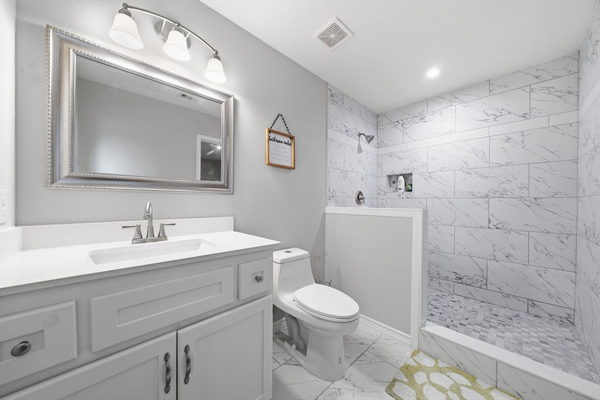 450 N Sunset Boulevard, Holiday Lakes, TX 77515 A bright, updated bathroom with a floor-to-ceiling marble-look walk-in shower, a built-in niche, and a mosaic tile floor. The quartz-topped vanity with white cabinetry and a three-light fixture keeps it clean, modern, and easy to maintain.