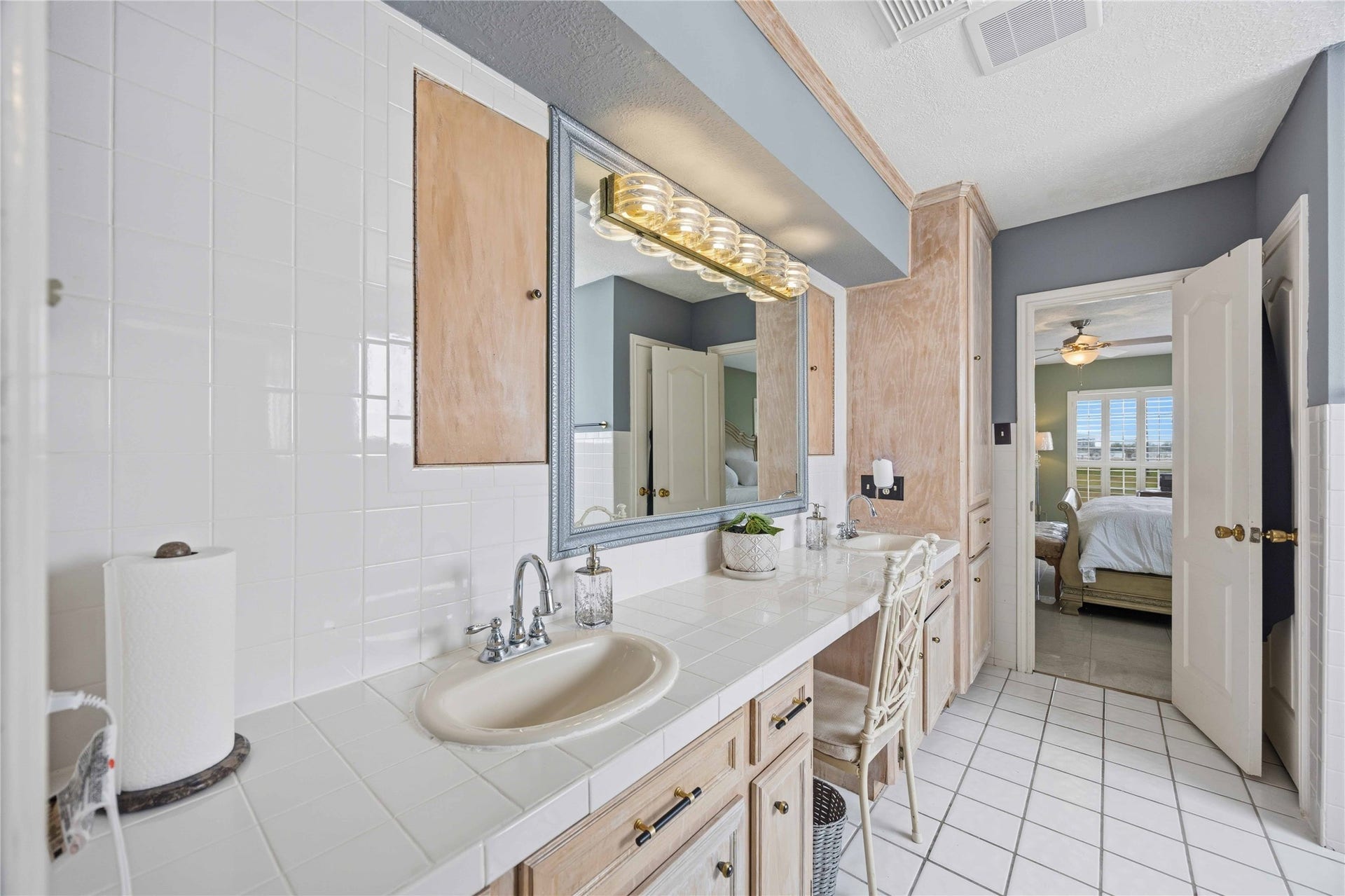 12075 FM 359 Road, #1, Hempstead, TX 77445 This bathroom features a spacious countertop with a single sink, ample cabinet storage, and a large mirror with overhead lighting. The neutral tile flooring complements the light wood cabinetry. A doorway leads to a bright, inviting bedroom with a ceiling fan and large window.