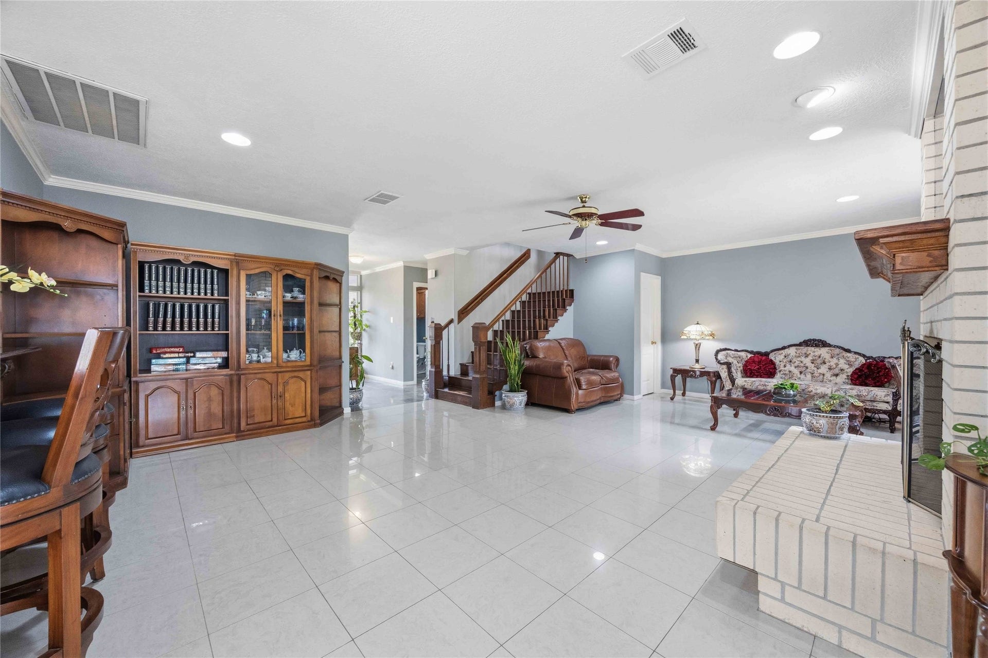 12075 FM 359 Road, #1, Hempstead, TX 77445 This spacious living room features glossy tile floors, a cozy fireplace, and elegant wood furnishings. It includes a built-in bookcase, a seating area, and access to an open staircase. The room is bright, with recessed lighting and large windows.