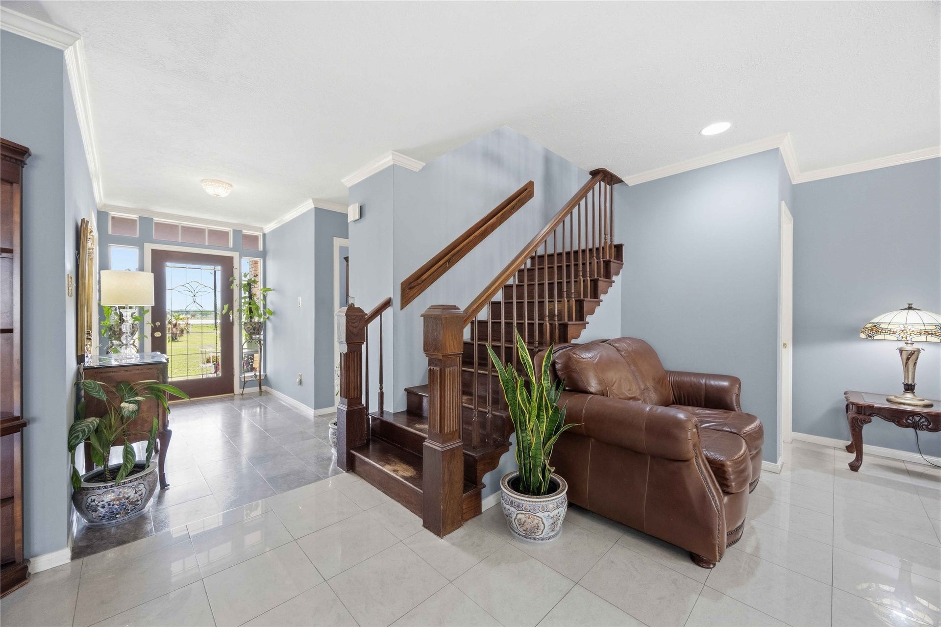 12075 FM 359 Road, #1, Hempstead, TX 77445 This photo showcases a bright entryway with a wooden staircase, tiled flooring, and neutral blue walls. The area is well-lit with natural light from a glass front door. There's a leather armchair and decorative plants, creating a welcoming atmosphere.