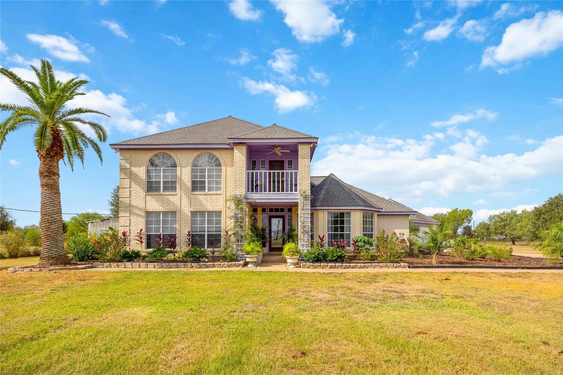 12075 FM 359 Road, #1, Hempstead, TX 77445 This photo showcases a charming two-story brick home with a landscaped front yard and a tall palm tree. The house features large arched windows, a welcoming front entrance, and a well-maintained lawn, set against a backdrop of a clear blue sky.