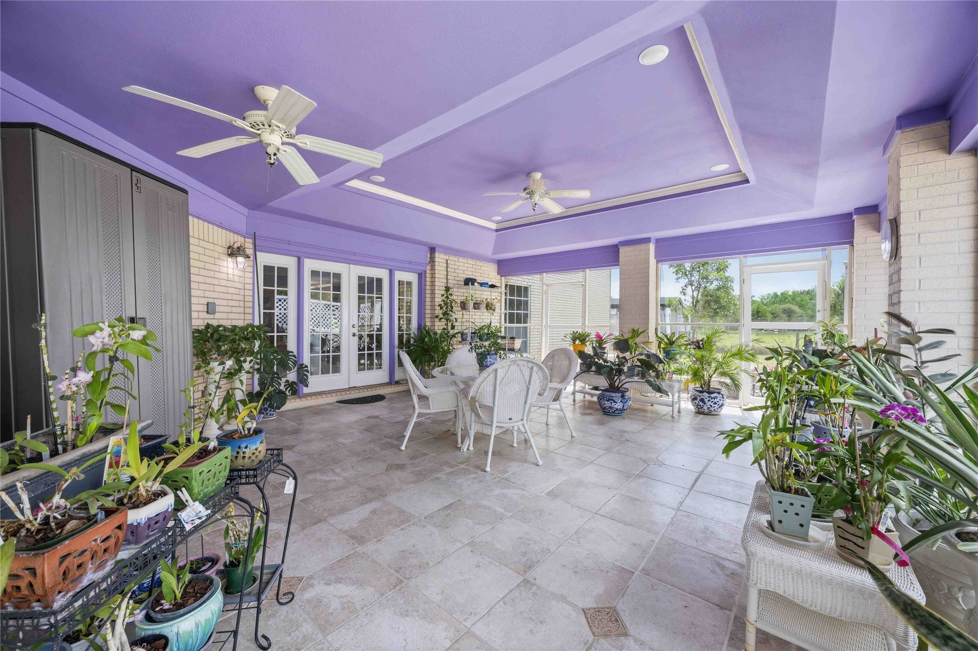 12075 FM 359 Road, #1, Hempstead, TX 77445 This spacious sunroom features a vibrant purple ceiling, large windows for natural light, and tile flooring. It's decorated with abundant plants and includes ceiling fans and comfortable seating, offering a perfect space for relaxation and entertaining.