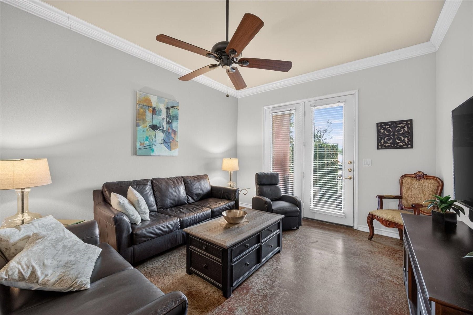 Charming living room with sleek leather seating, a ceiling fan, and abundant natural light streaming through large windows, creating a cozy and inviting atmosphere.