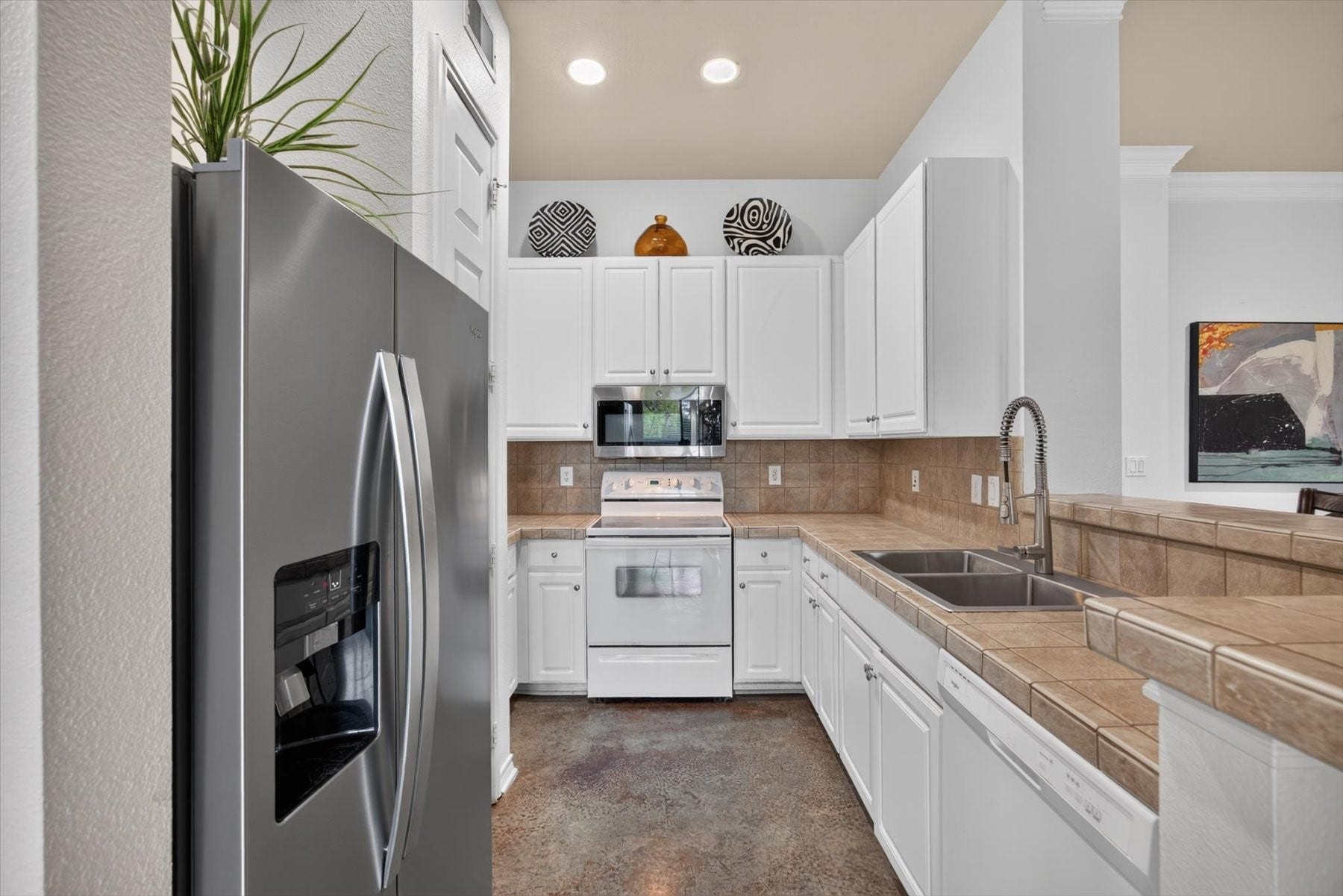 Modern kitchen featuring sleek stainless steel refrigerator, double sink, white cabinetry, and a practical tiled countertop.