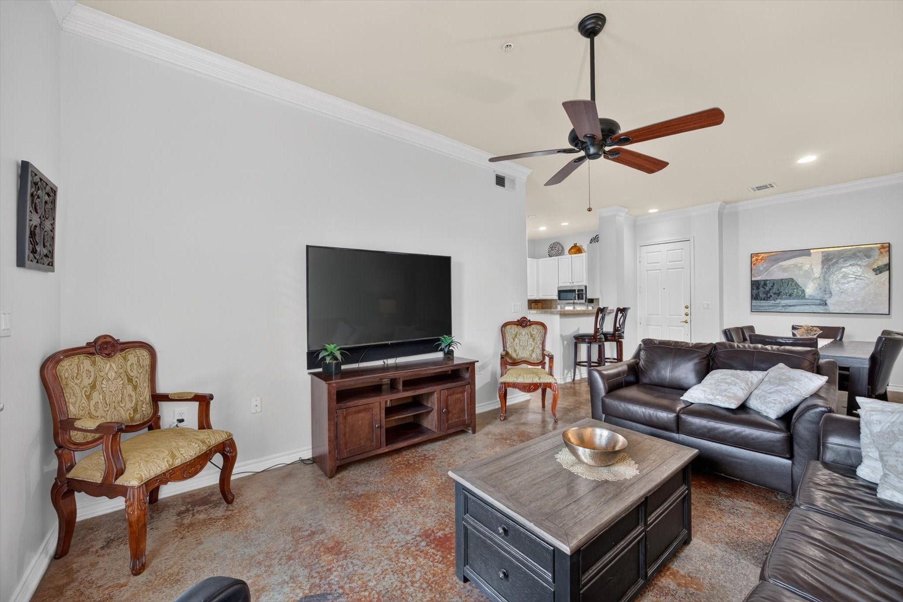 Spacious living area with elegant crown molding, polished flooring, and a large ceiling fan. The open layout features a comfortable seating arrangement, a wall-mounted TV, and a seamless flow into the dining and kitchen areas, perfect for entertaining.