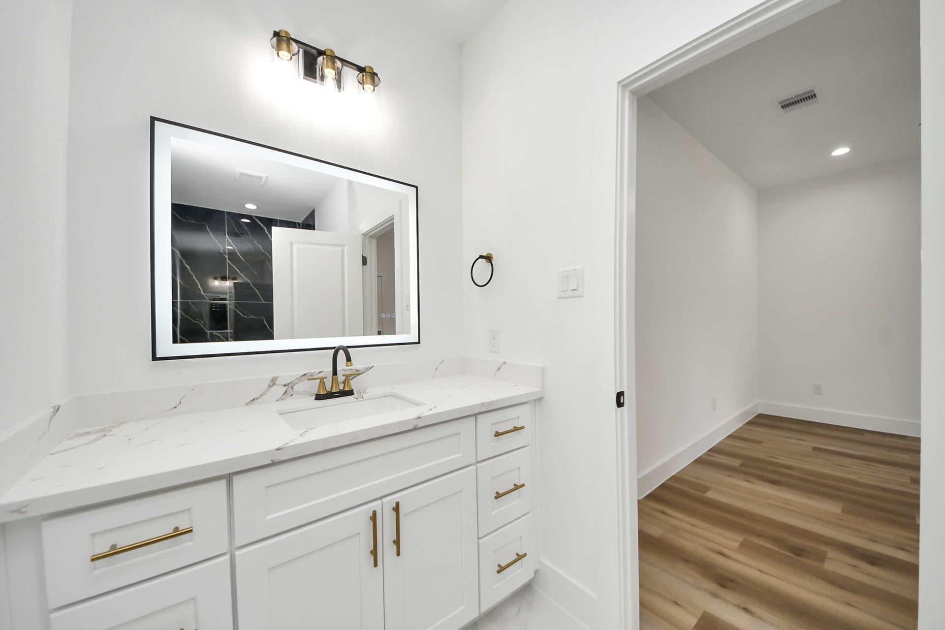 7801 King Street, Houston, TX 77028 Elegant bathroom featuring a sleek vanity with a marble countertop, modern fixtures, and a large illuminated mirror, leading to a room with stylish wood flooring.