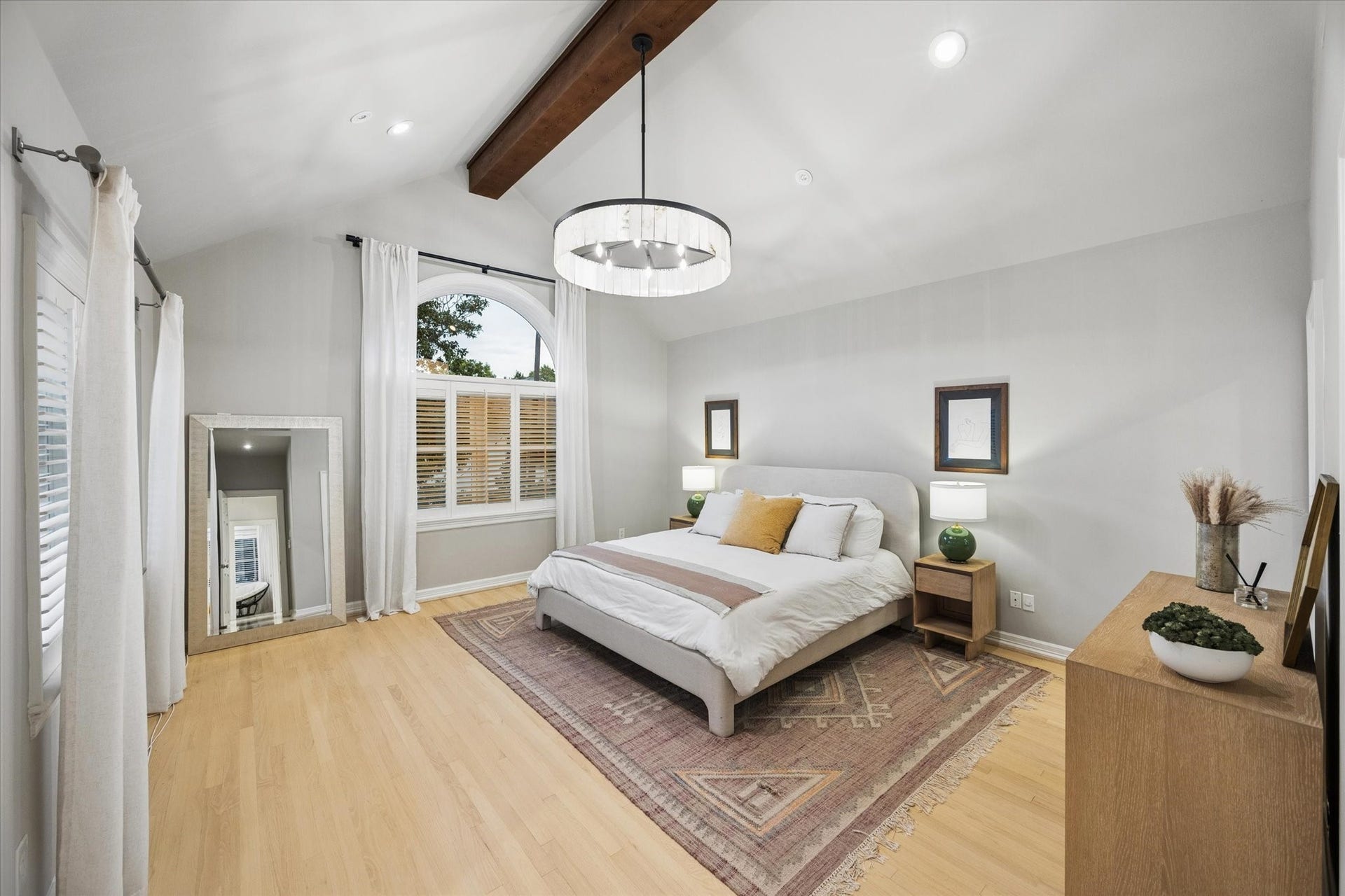 Spacious primary suite with a dramatic vaulted ceiling, exposed wood beam, beautiful hardwood floors and a spectacular oversized arched window that fills the room with natural light and adds striking architectural character.