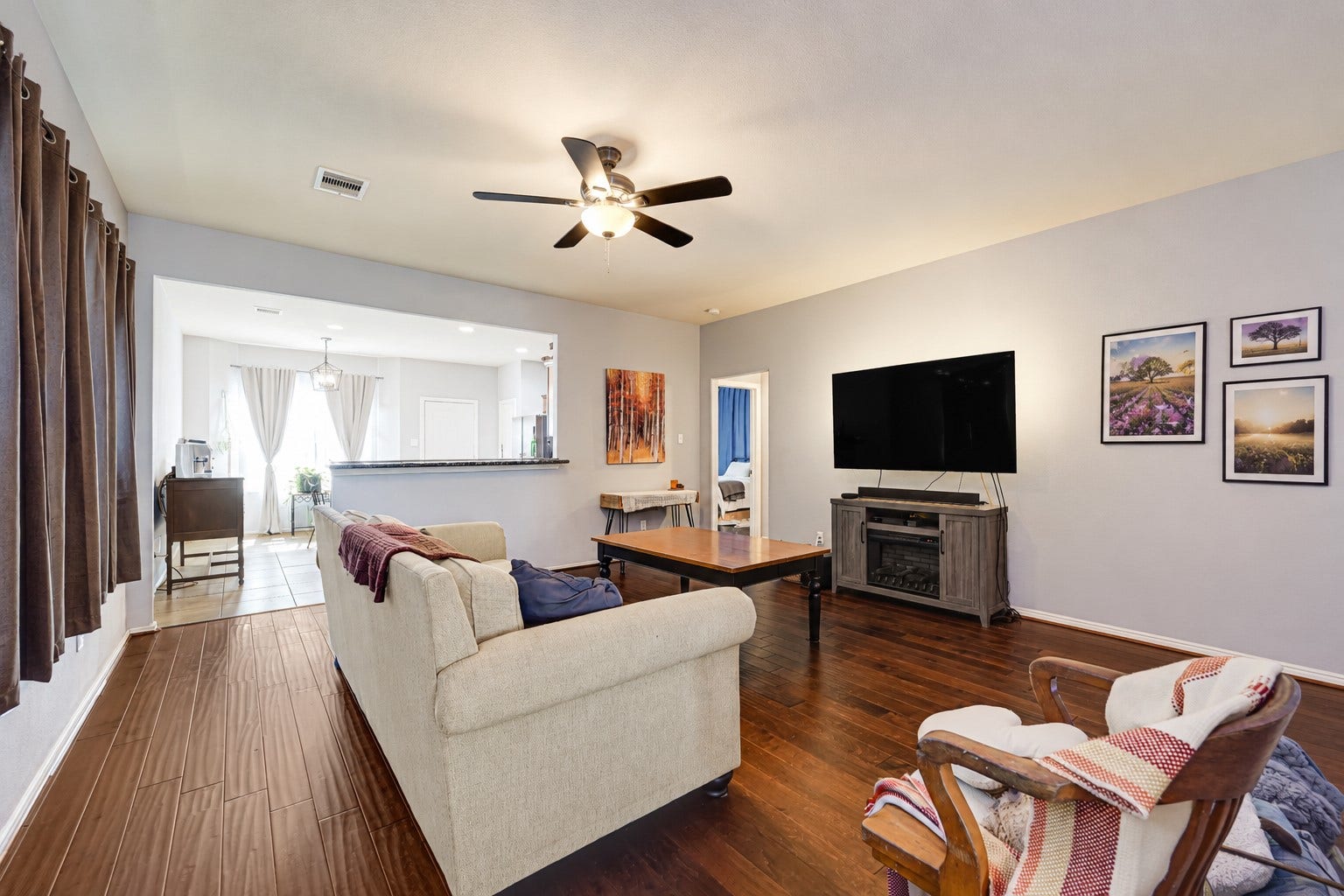 Spacious living room featuring rich hardwood floors, a cozy fireplace, and a large wall-mounted TV, seamlessly flowing into a bright dining area with elegant drapes.