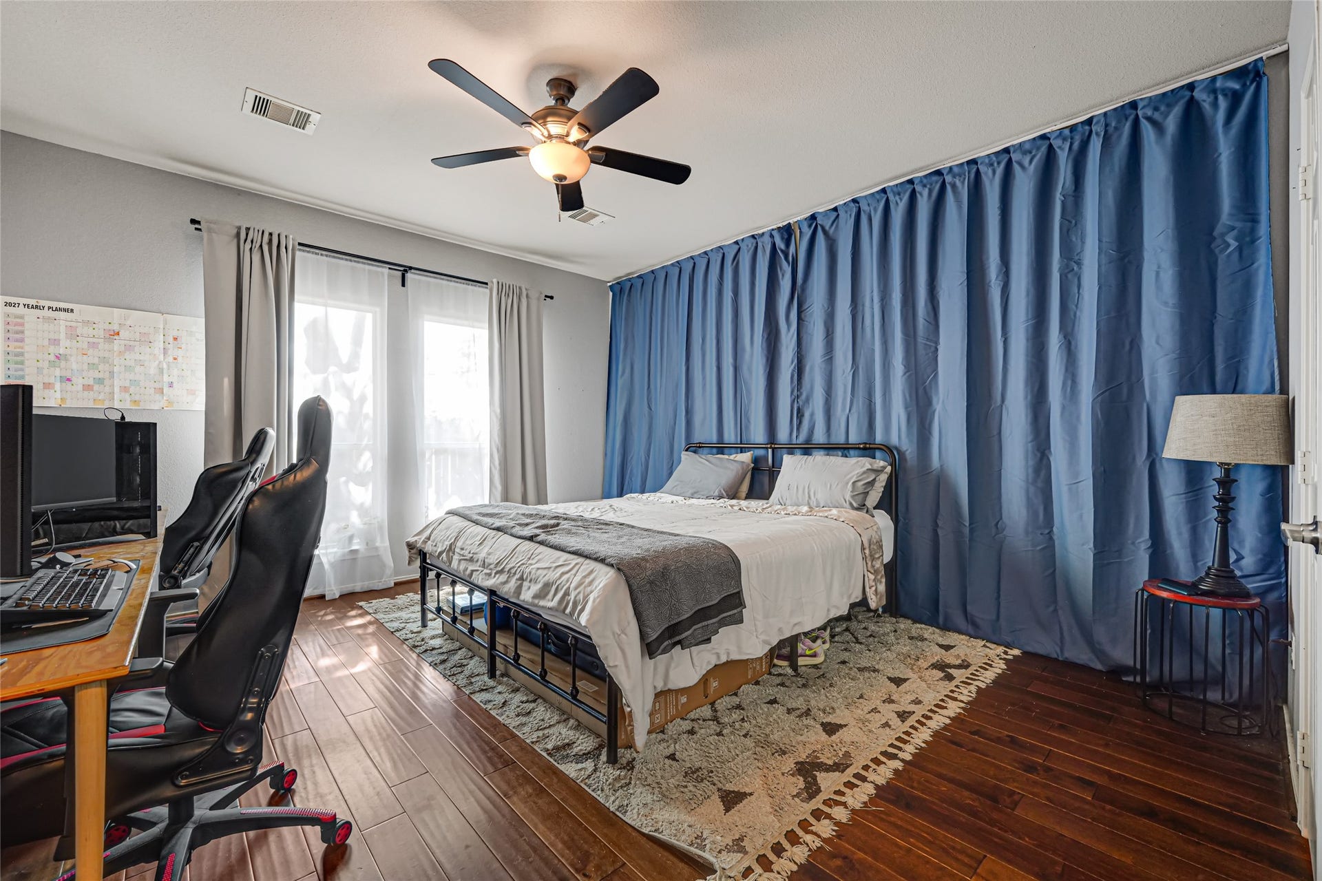 Spacious bedroom with rich hardwood floors, featuring a cozy bed, stylish blue curtains, and a practical workspace area.