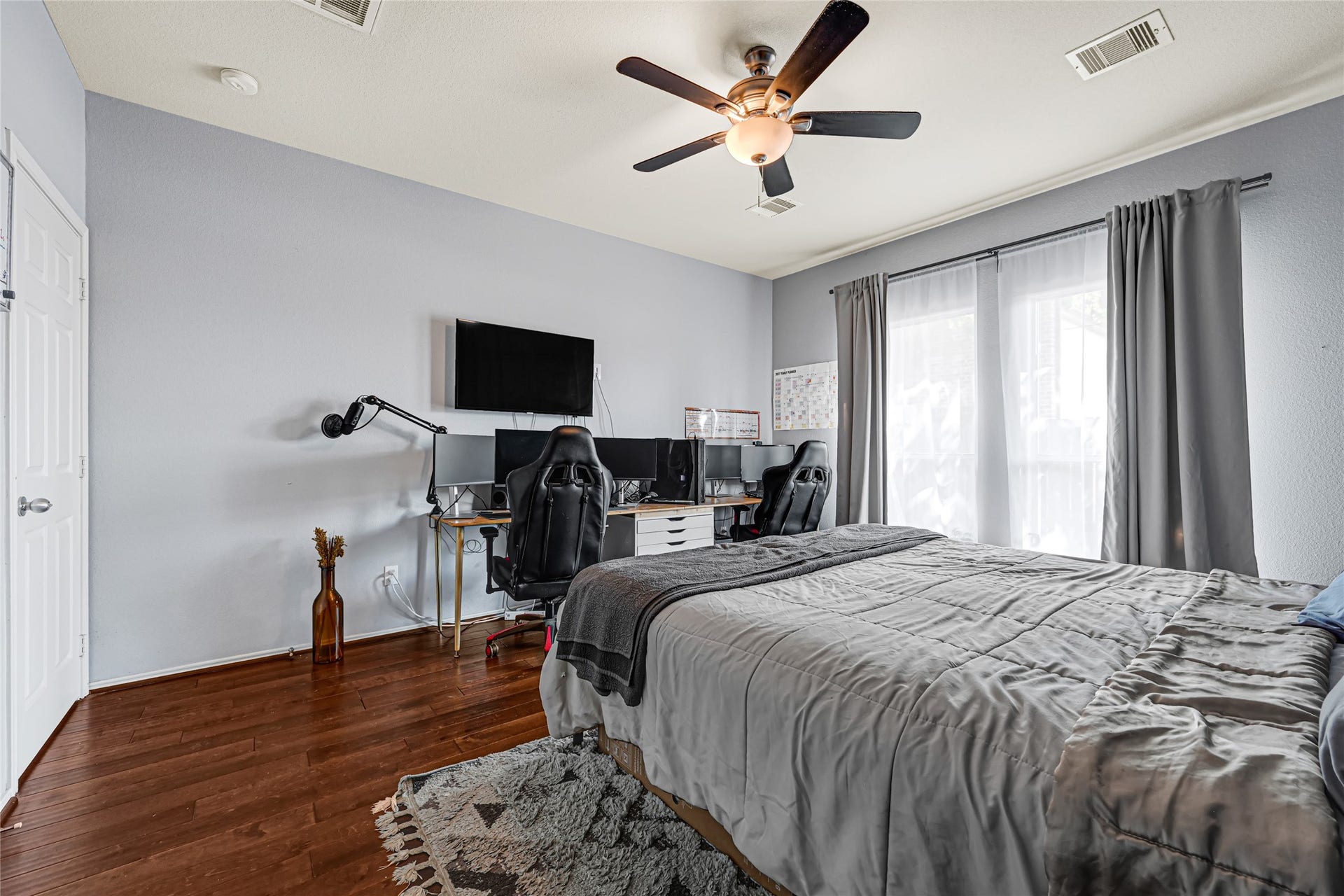 Spacious bedroom with warm wood flooring, a modern ceiling fan, and ample natural light from large windows. Features a cozy bed and a functional workspace with multiple screens, perfect for relaxation and productivity.