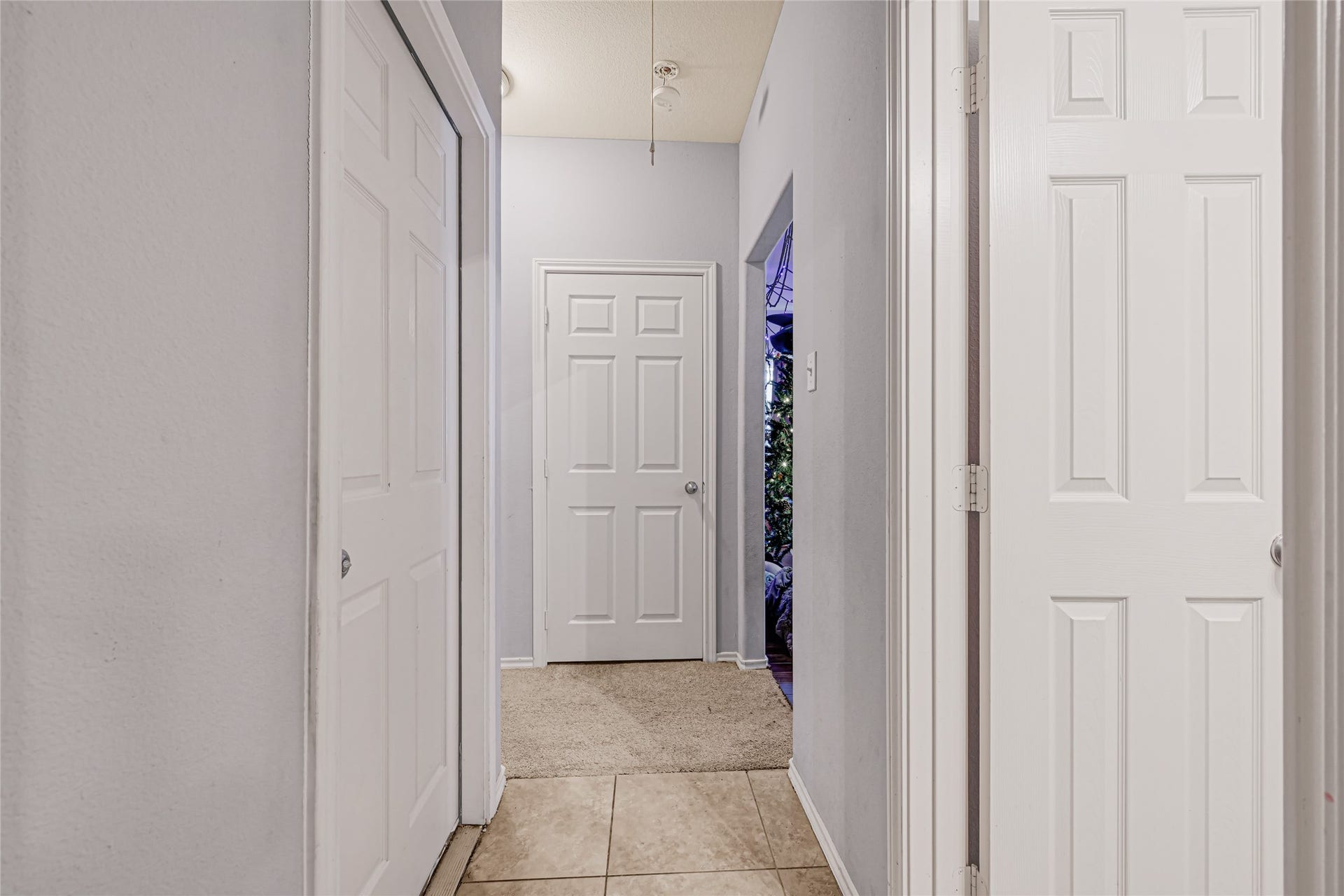 Bright hallway with neutral tones, featuring multiple doors and tile flooring transitioning to carpet, leading to a cozy space.