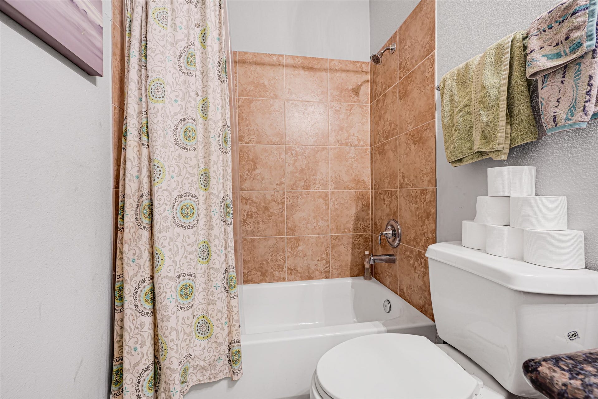Charming bathroom with a cozy tub and warm-toned tile surround, complemented by a patterned shower curtain and convenient shelving space above the toilet.
