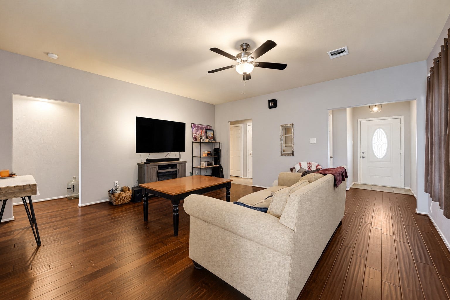 Spacious living room with warm hardwood floors, a cozy fireplace, and ample seating. The modern ceiling fan and large TV create a perfect space for relaxation and entertainment.