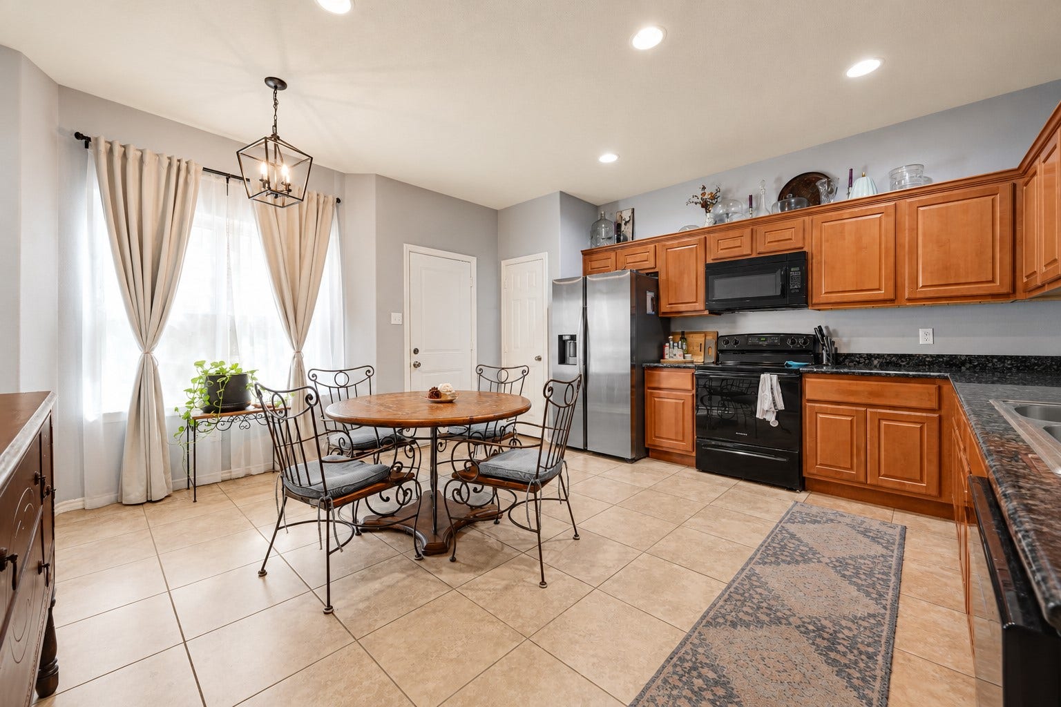 A spacious kitchen with warm wood cabinetry, granite countertops, and modern appliances, complemented by a cozy dining area with elegant lighting and natural light from large windows.