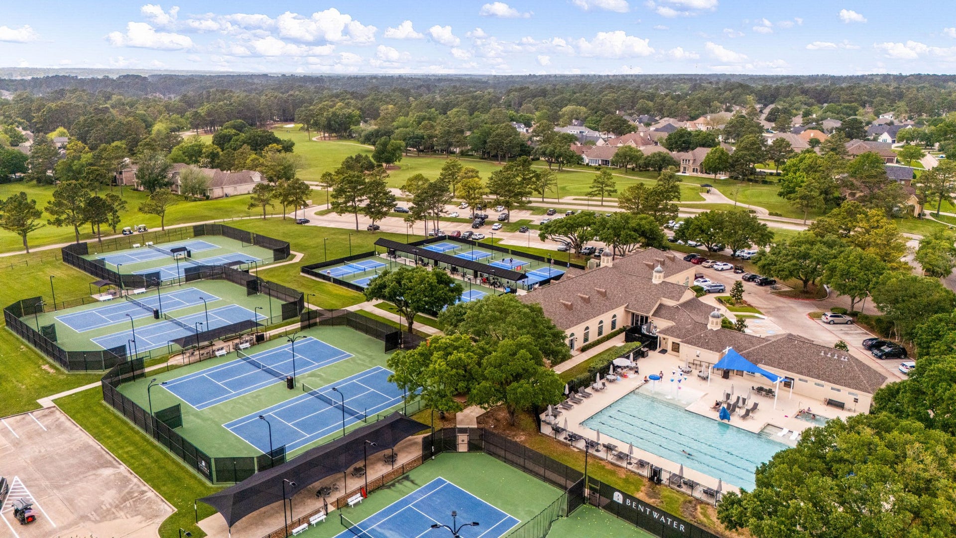 98 Camden Hills Drive, Montgomery, TX 77356 This photo showcases a vibrant community setting featuring multiple tennis courts, a large swimming pool, and a clubhouse. The area is surrounded by lush greenery and residential homes, offering recreational amenities in a serene neighborhood environment.