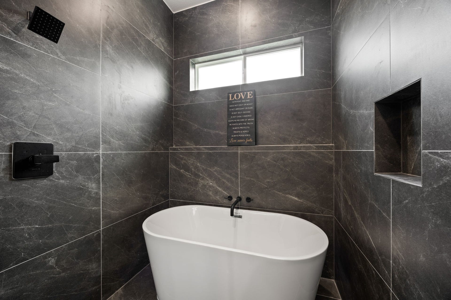 98 Camden Hills Drive, Montgomery, TX 77356 This bathroom features a sleek, modern design with dark marble tiles, a freestanding white bathtub, and a built-in wall niche for storage. A large window provides natural light, creating a serene atmosphere.
