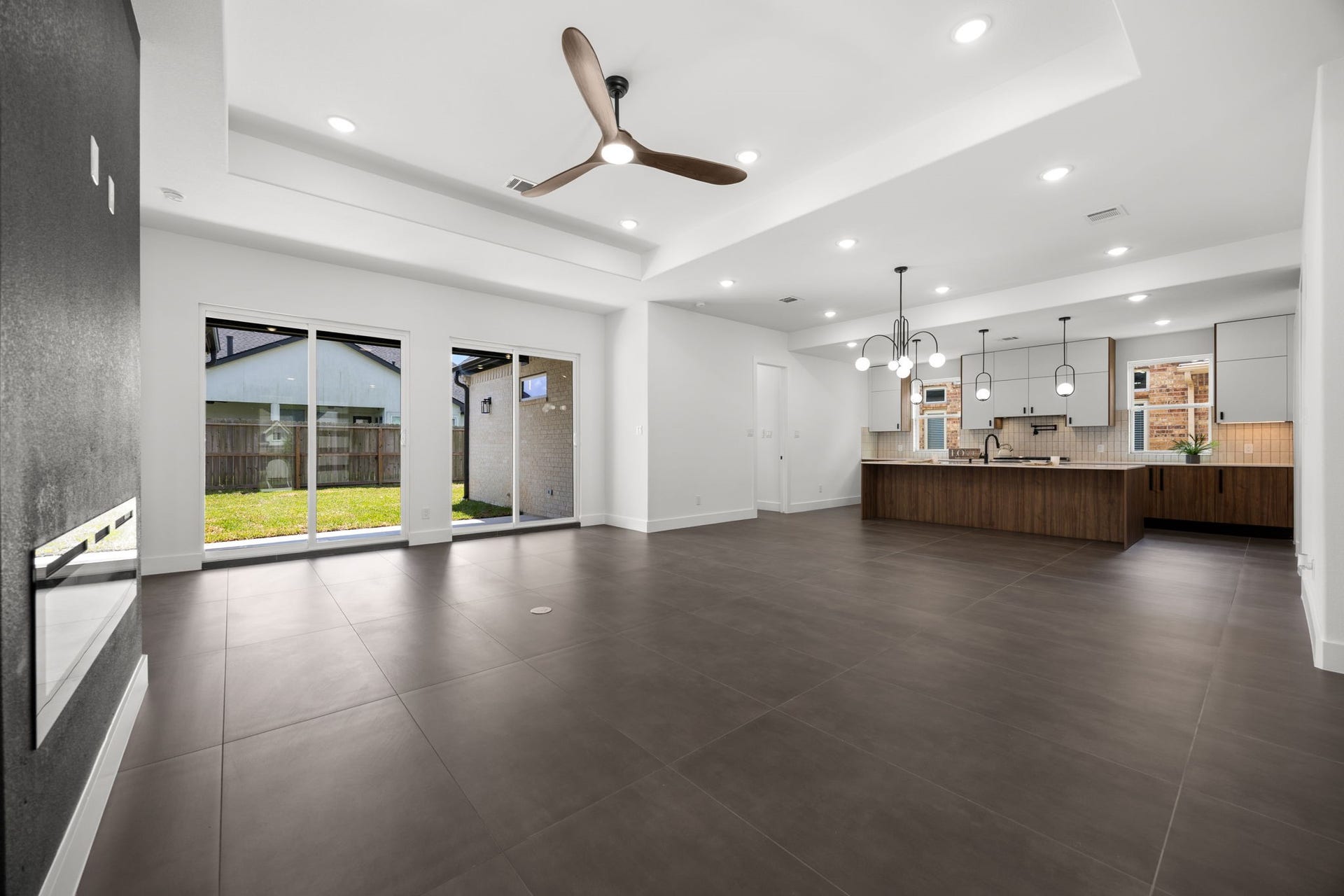 98 Camden Hills Drive, Montgomery, TX 77356 This photo showcases a spacious open-concept living area with modern design elements. It features large tile flooring, recessed lighting, and a ceiling fan. The room seamlessly connects to a stylish kitchen with a large island, contemporary pendant lights, and ample natural light from sliding glass doors leading to a fenced backyard.