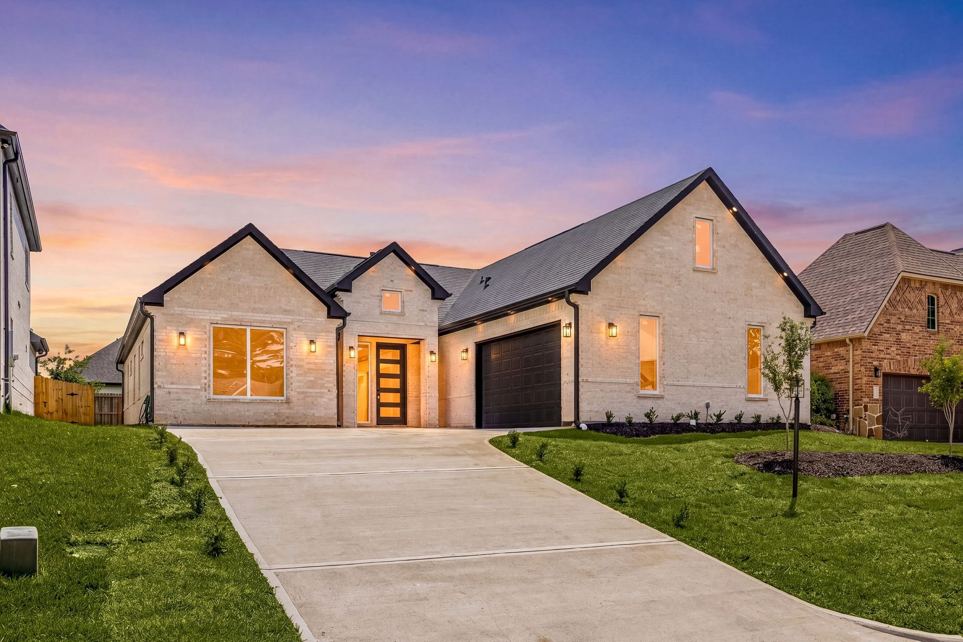 98 Camden Hills Drive, Montgomery, TX 77356 This photo showcases a modern single-story home with a clean, light brick facade and dark roof accents. It features a spacious driveway leading to a two-car garage and a well-maintained front lawn. The house is warmly lit, enhancing its curb appeal against a colorful sunset sky.