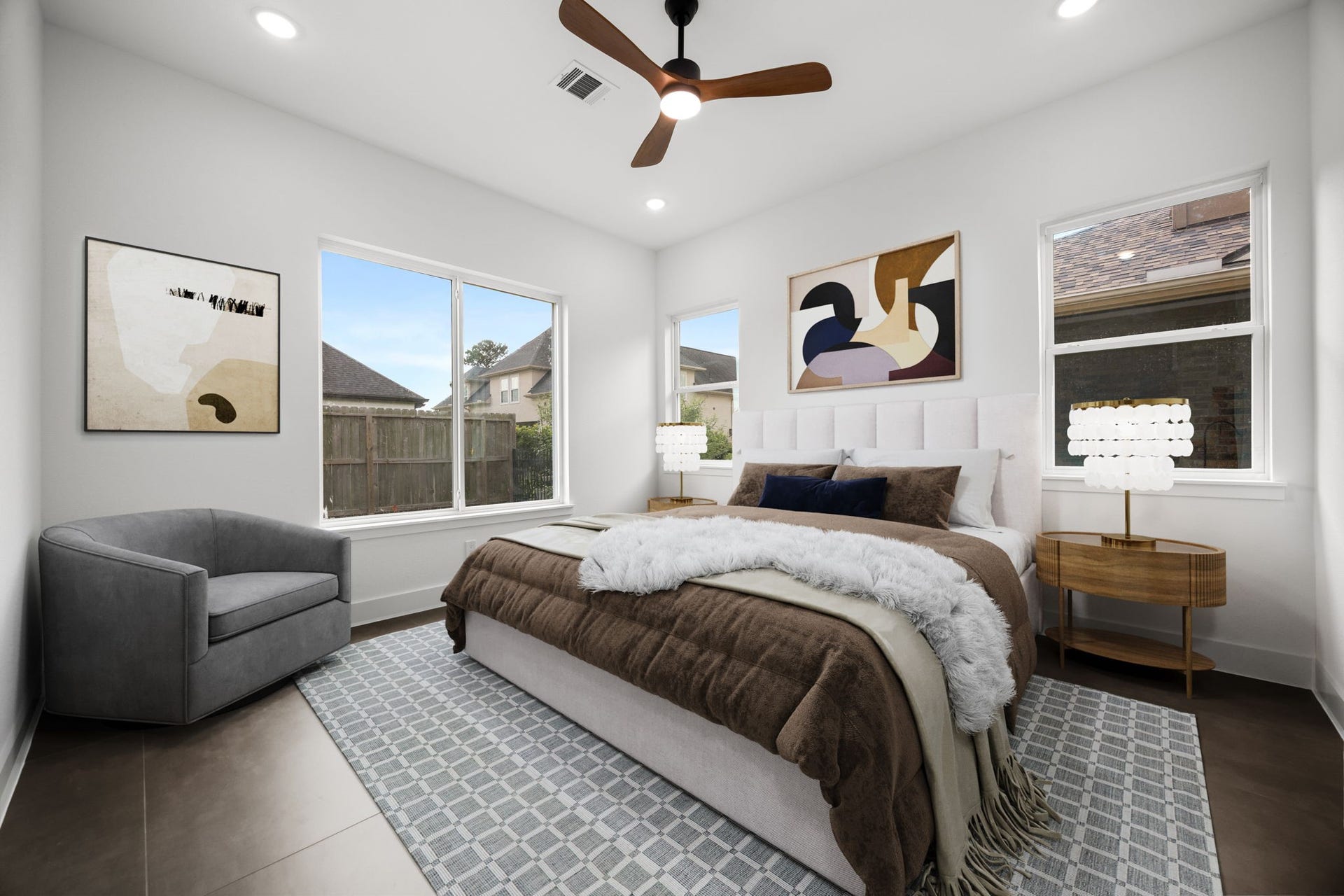 98 Camden Hills Drive, Montgomery, TX 77356 This bedroom features a modern design with a cozy bed, abstract artwork, and ample natural light from two large windows. The space includes elegant side tables, a plush gray armchair, and a ceiling fan, creating a comfortable and stylish retreat.