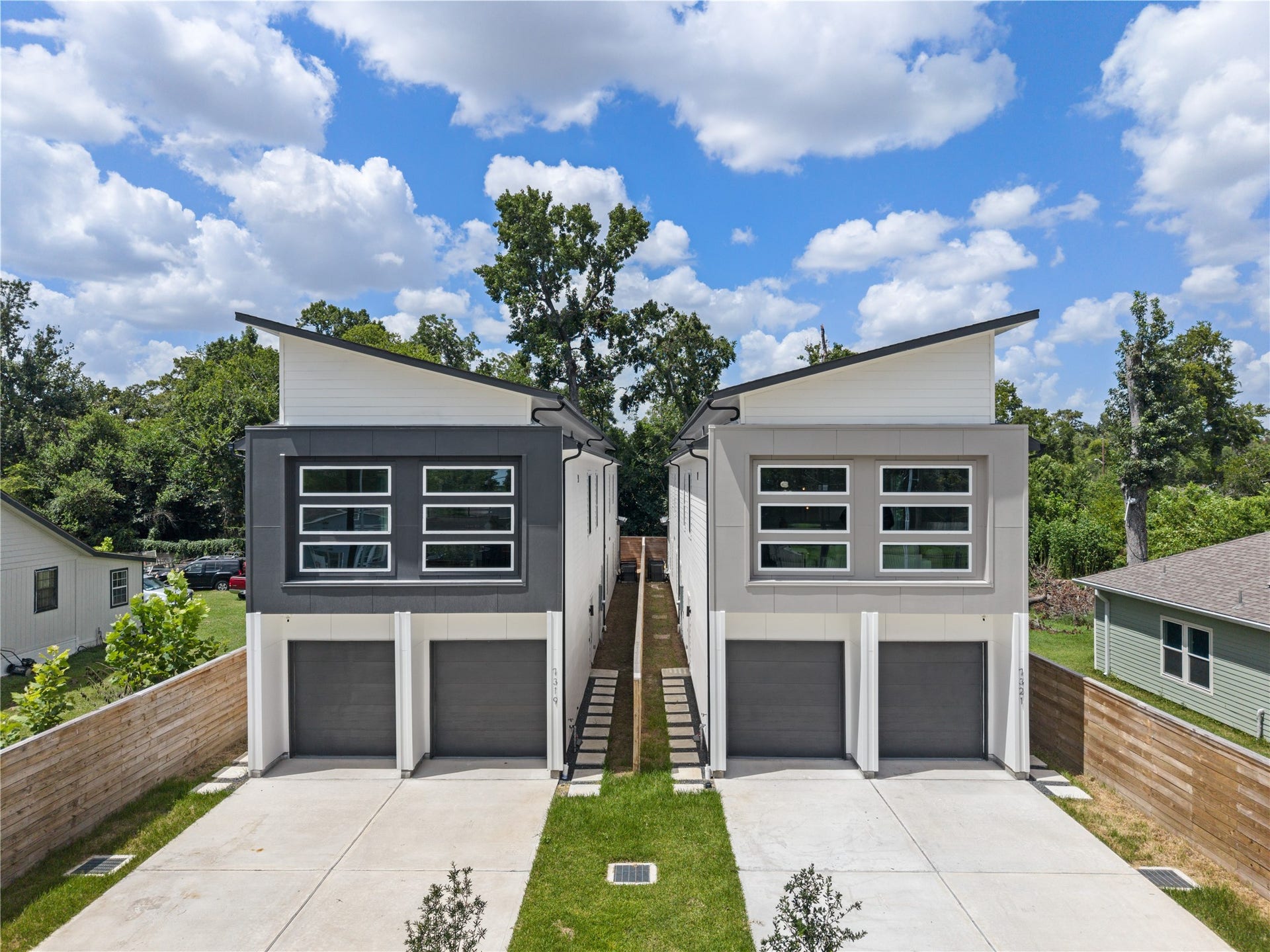 Modern duplex featuring sleek, angular rooflines and large windows for ample natural light. Eachunit offers a private garage and a shared driveway, surrounded by a lush, tree-lined setting