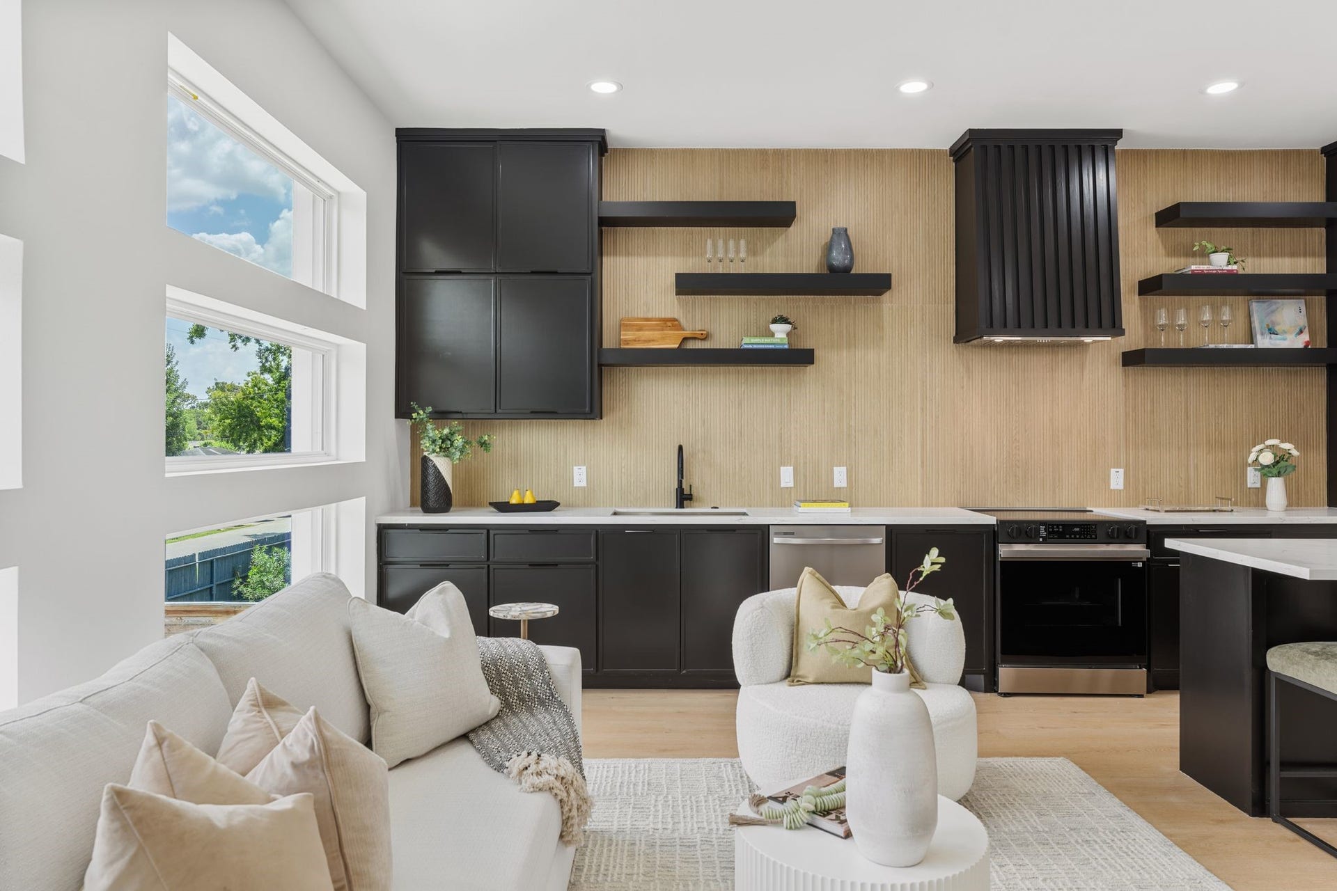 Stunning modern kitchen with sleek black cabinetry, open shelving, and a cozy adjacent living area.Ample natural light pours in through unique geometric windows, accentuating the elegant design.