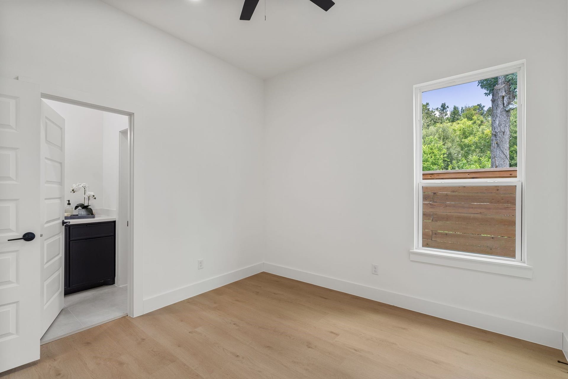 Bright, airy room with sleek hardwood floors and amodern ceiling fan, featuring a window with lush green views and an adjacent bathroom with contemporary fixtures.