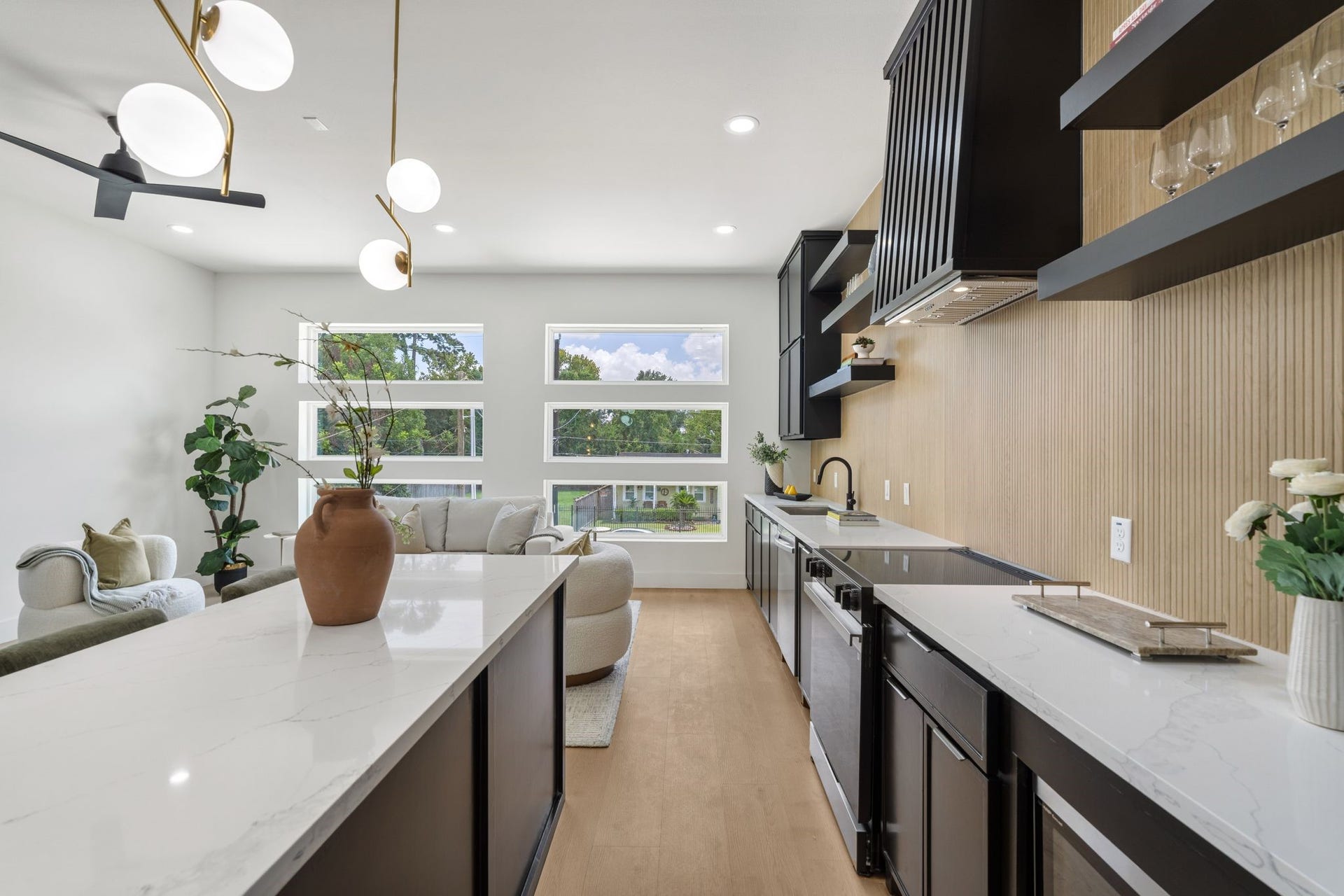 Sleek and modern kitchen with elegant black cabinetry, spacious white countertops, and stylish open shelving. Large windows fill the space with natural light, complementing the contemporary light fixtures and warm wood accents.