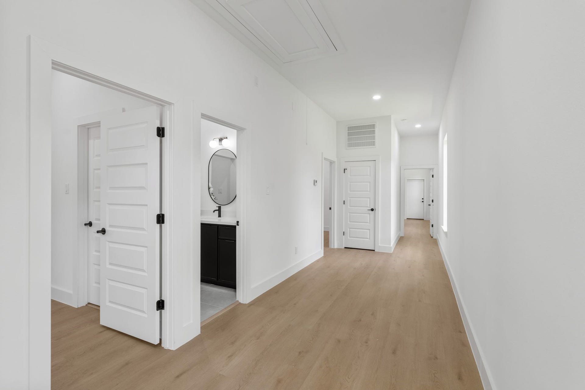 Bright, spacious hallway with elegant wood flooring, modern light fixtures, and crisp white walls leading to multiple rooms, including a stylish bathroom.