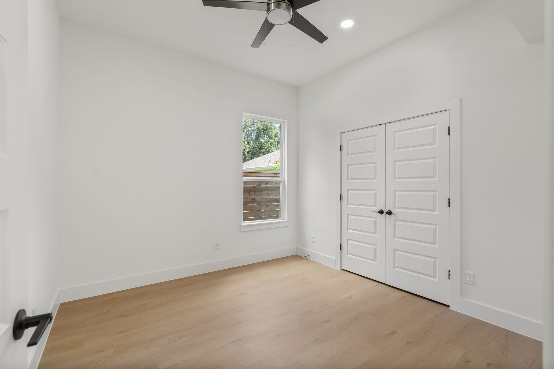 Bright and airy room featuring sleek hardwood floors, a modern ceiling fan, and ample natural light from a single window, complemented by double doors for added storage.