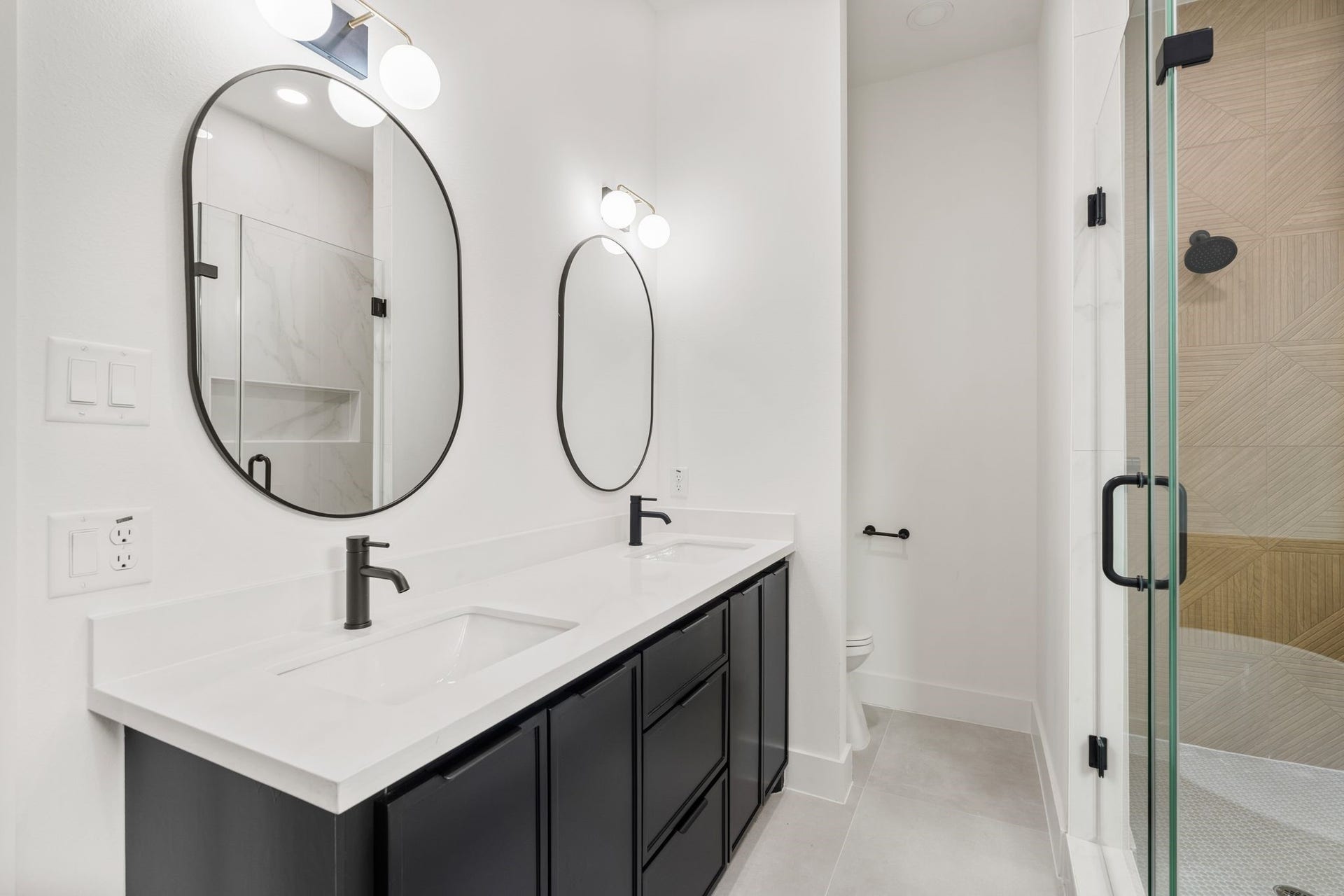 Elegant bathroom featuring a modern double vanity with sleek black fixtures, oval mirrors, and a glass-enclosed shower.