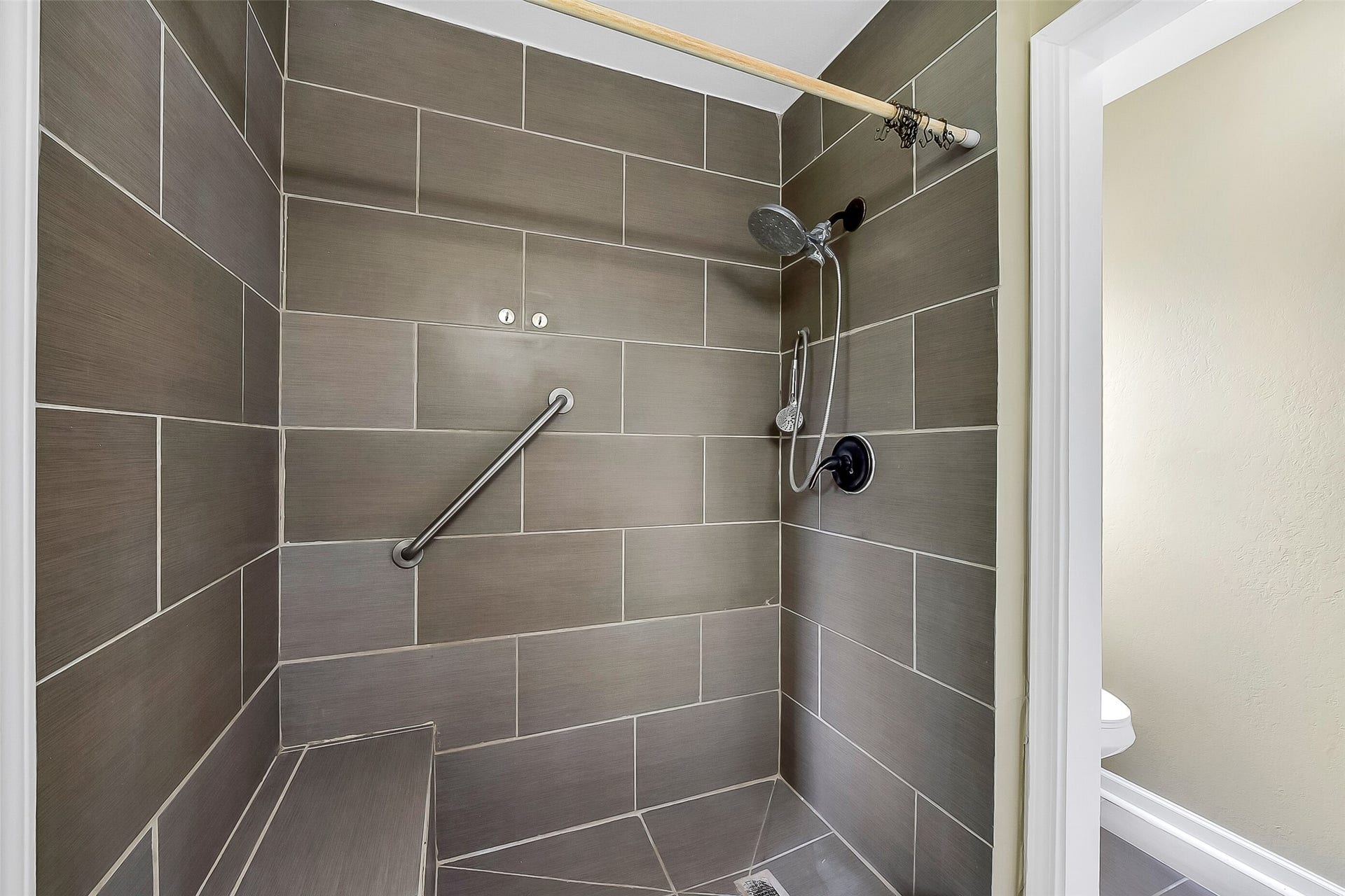 15503 Wandering Trail, Friendswood, TX 77546 The master bathroom features a modern design with a sleek vanity topped with a solid-surface countertop, framed mirror, and updated lighting. The spacious walk-in shower is accented with oversized tile, a built-in bench, a grab bar, and dual showerheads, including a rain-style fixture.