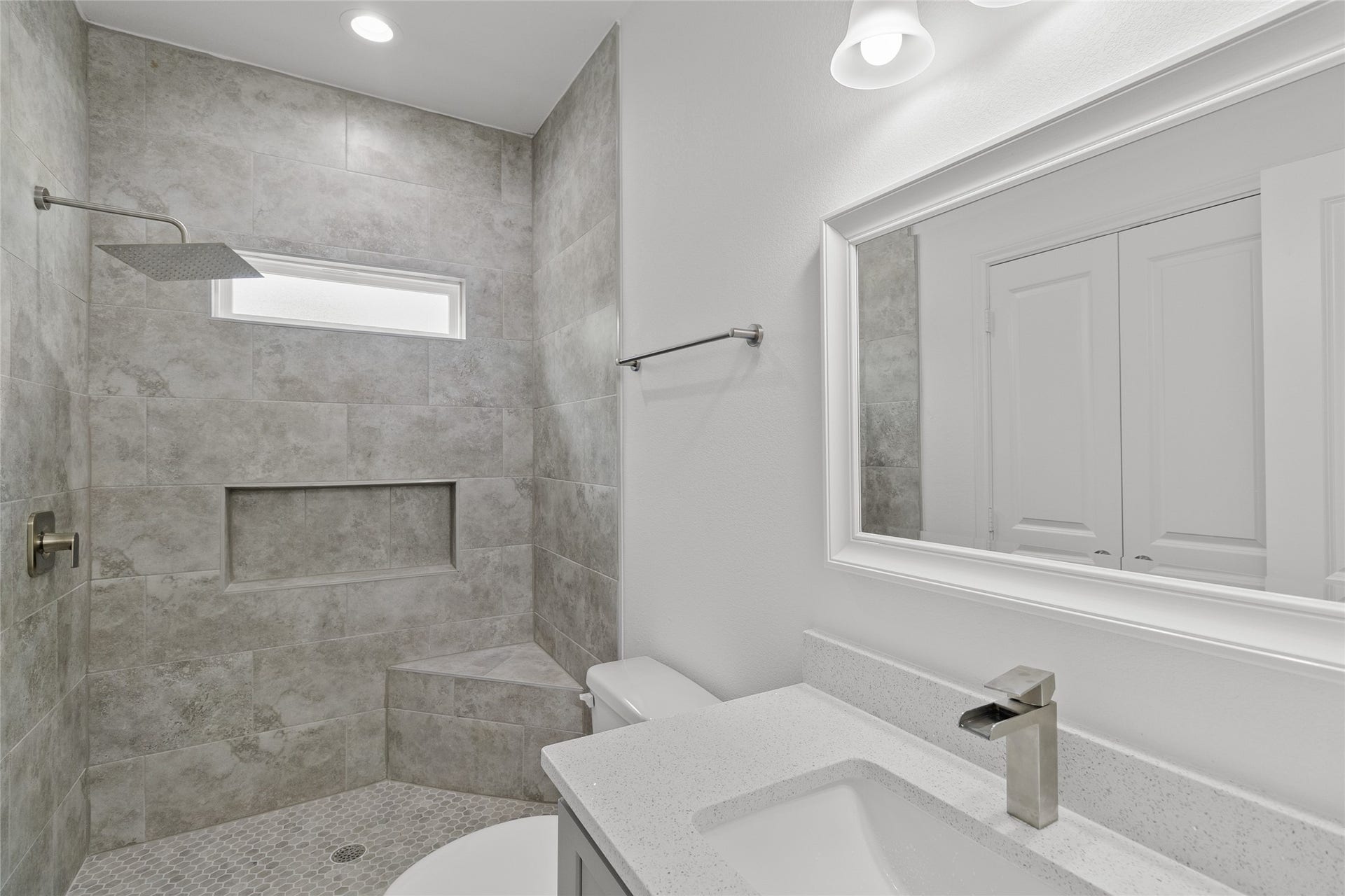 760 Juneau Court, Montgomery, TX 77316 Modern bathroom featuring a large walk-in shower with gray tile, a built-in niche for toiletries, and a sleek rain shower head. The vanity includes a spacious countertop with an undermount sink and a large mirror with white trim. A small window provides natural light.