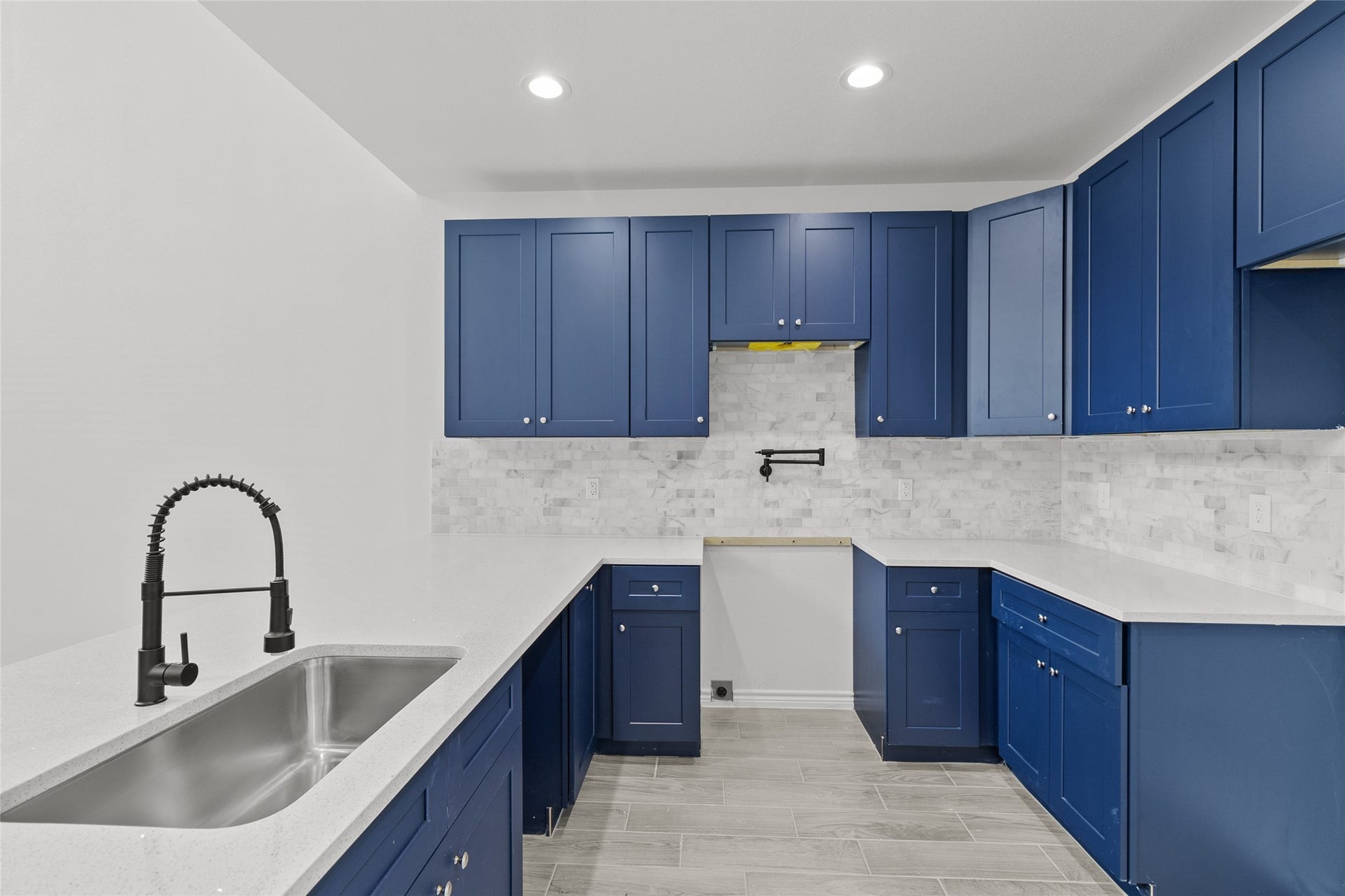 760 Juneau Court, Montgomery, TX 77316 This is a modern kitchen featuring sleek blue cabinetry, gray tile backsplash, and light countertops, complete with a deep sink and a high-arc faucet. The room has recessed lighting and gray flooring, creating a contemporary and stylish space.