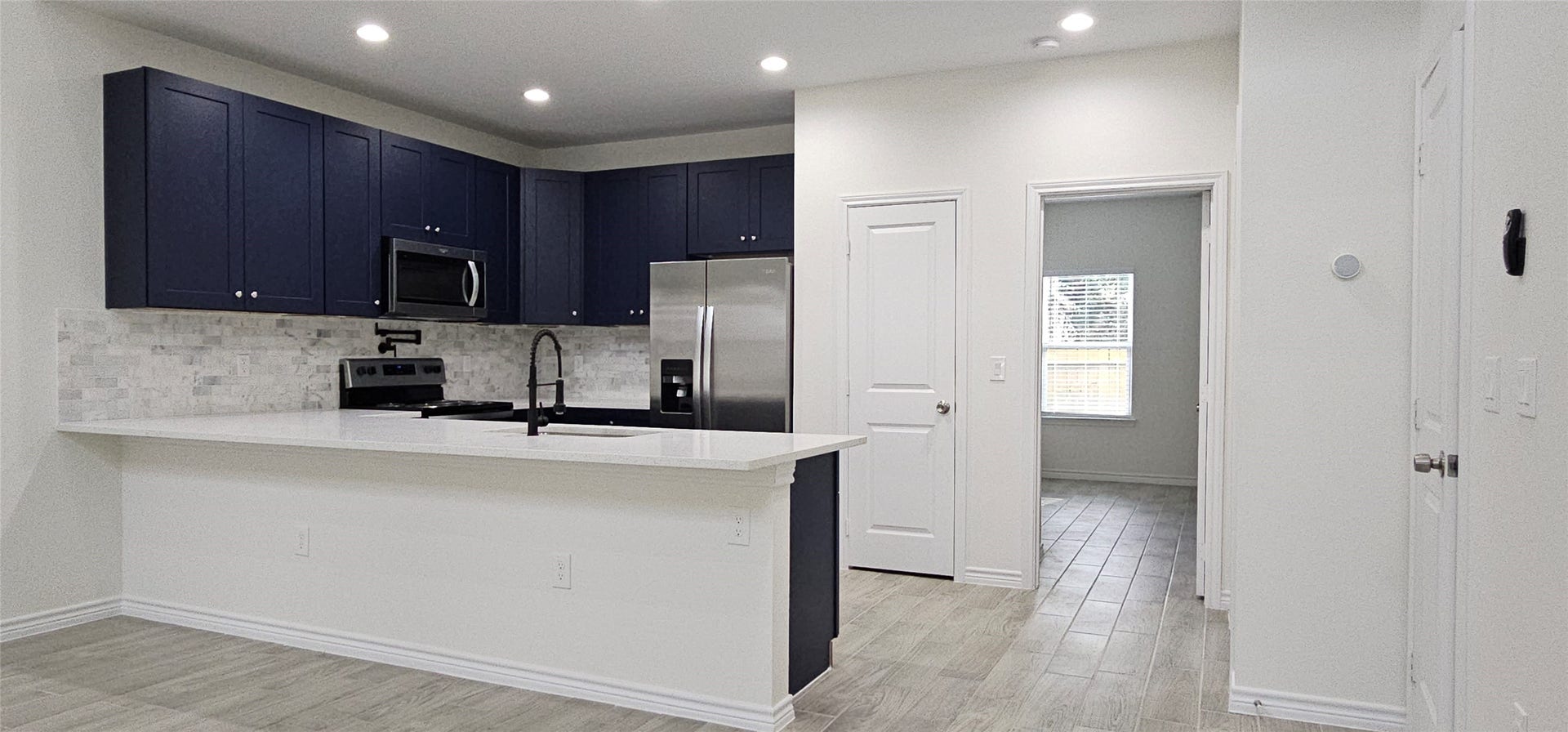 760 Juneau Court, Montgomery, TX 77316 This is a modern kitchen with dark blue cabinetry, stainless steel appliances, and a white subway tile backsplash. It features a central island with a sink and room for seating, complemented by light wood-look flooring and neutral wall colors. A doorway leads to an adjacent room with a window for natural light.