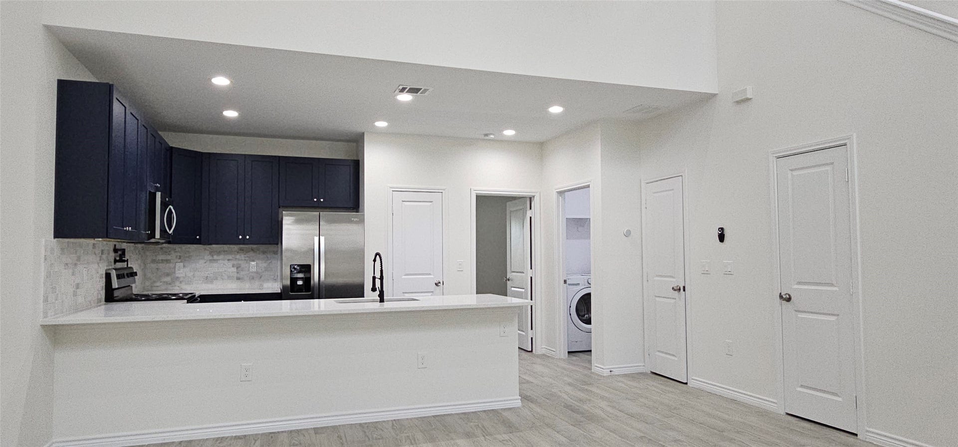 760 Juneau Court, Montgomery, TX 77316 This is a modern, open-concept living space featuring a kitchen with dark cabinets and stainless steel appliances, a breakfast bar, recessed lighting, and a view into a laundry room. The area is finished with light walls and wood-like flooring for a clean, airy feel.