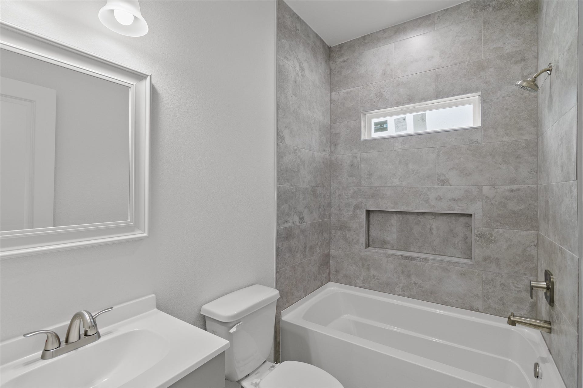 760 Juneau Court, Montgomery, TX 77316 This is a modern bathroom featuring a large mirror, under-lit vanity with a sink, toilet, and a full bathtub with gray tile surround. There's also a small frosted window providing natural light.