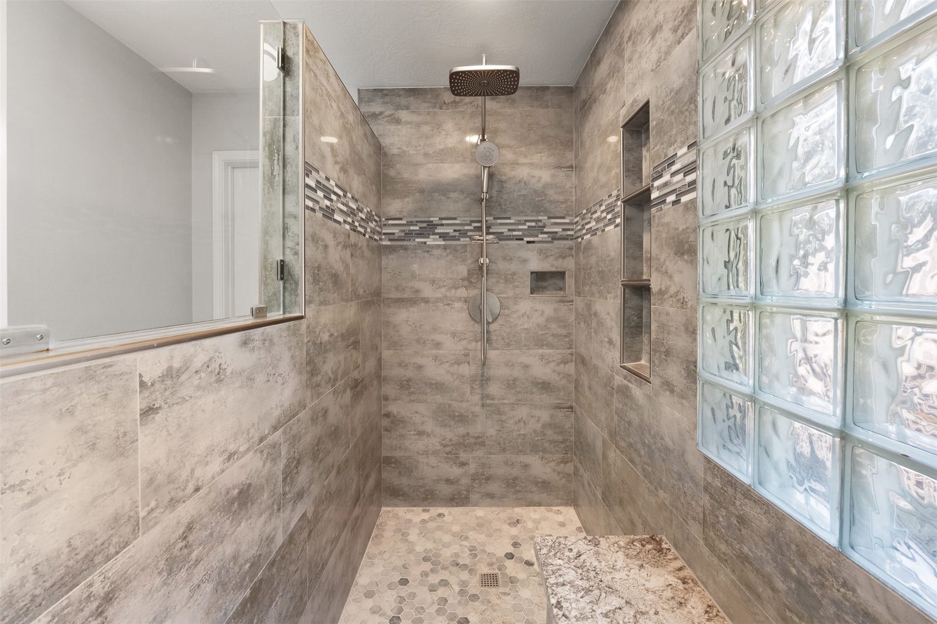 1118 Wentworth Drive, Pearland, TX 77584 Lined with large-format grey tile, this renovated shower features a rain shower head, glass block window, built-in bench, and hexagon tile floor with a mosaic accent band.