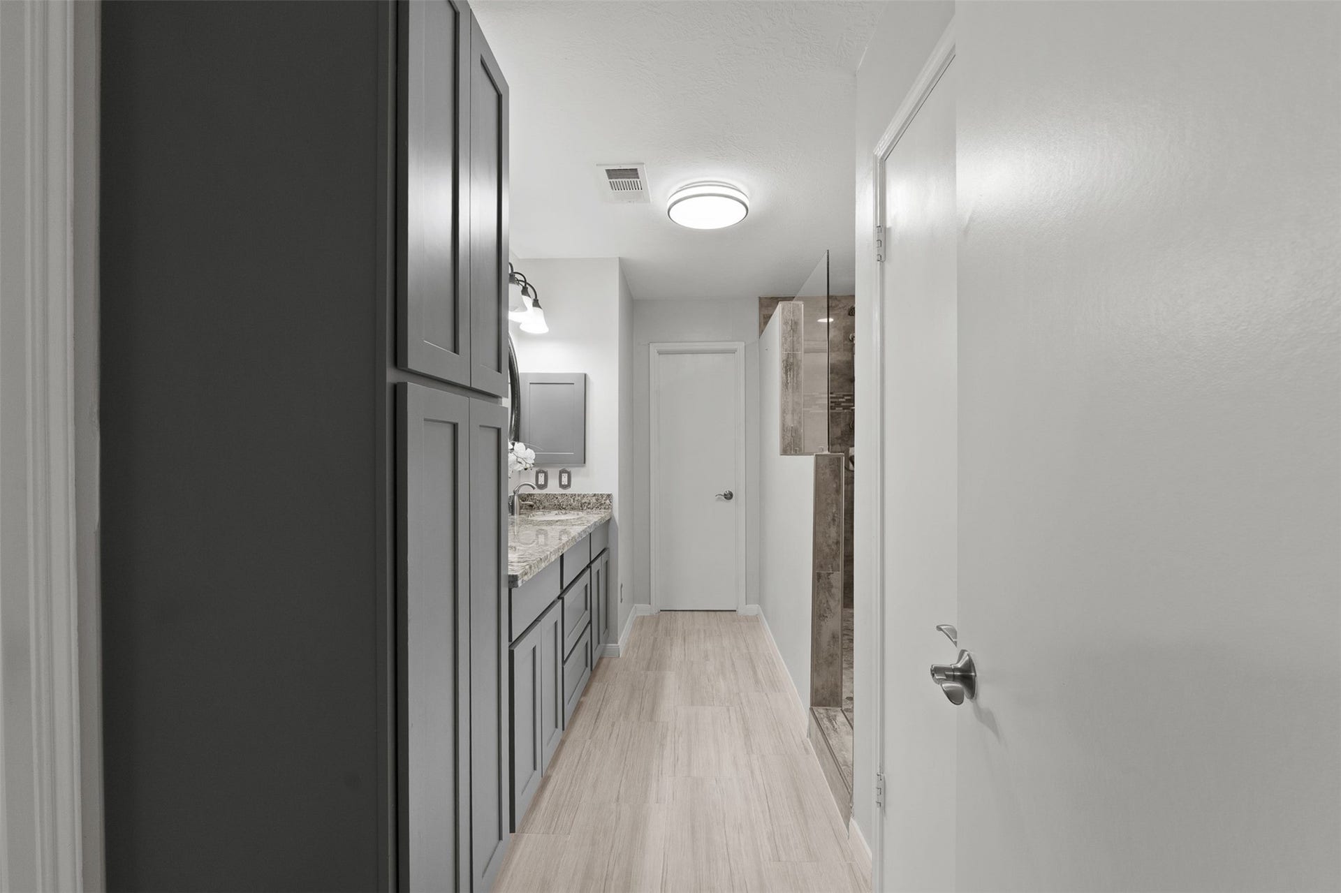 1118 Wentworth Drive, Pearland, TX 77584 Custom tall grey cabinetry lines the hall of this renovated primary bath, offering abundant storage space alongside wood-look tile flooring. Modern vanity with granite counters features a dual sink, complemented by a sleek rain shower with glass enclosure.