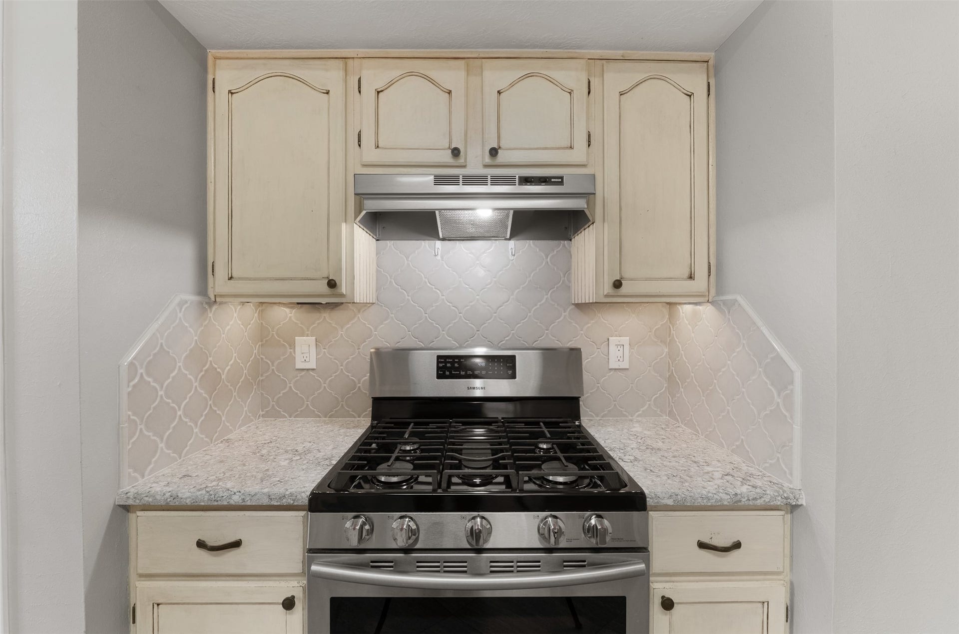 1118 Wentworth Drive, Pearland, TX 77584 Featuring a gas range with an updated range hood, this kitchen highlight showcases an intricate arabesque designer backsplash flanked by distressed cream cabinetry. Under-cabinet lighting illuminates the quartz countertops, providing both functionality and style.