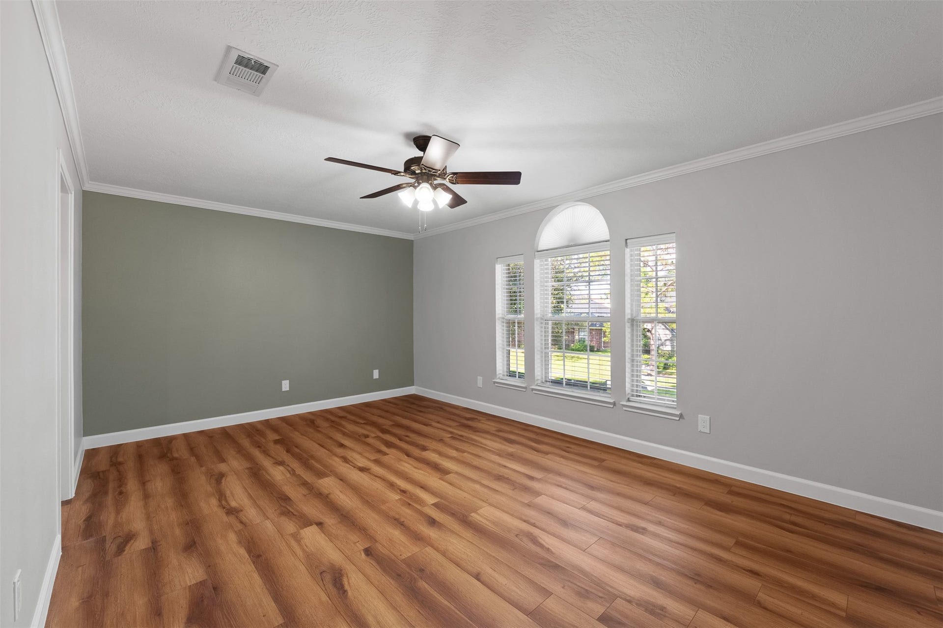 1118 Wentworth Drive, Pearland, TX 77584 Warm wood-look flooring stretches across this light-filled room, framed by crisp crown molding and a sage green accent wall. Wide windows with a center arch fill the space with light, while a dark wood ceiling fan provides steady breeze and brightness.