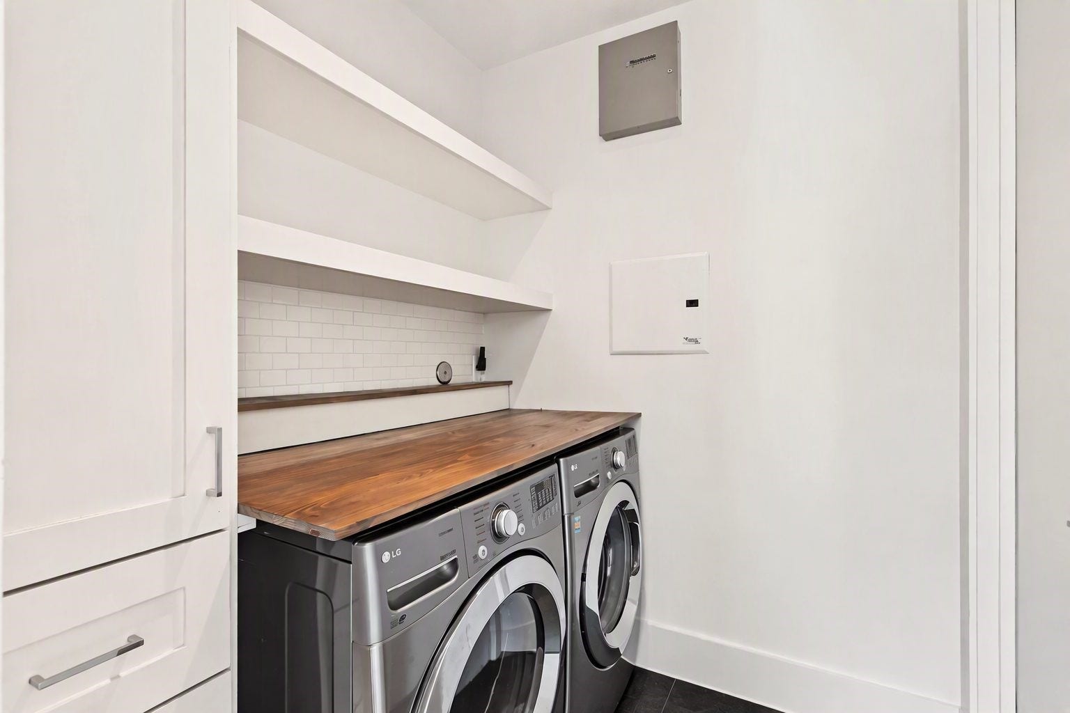 This dedicated laundry space effortlessly blends daily utility with modern design, pairing crisp white built-in cabinetry with dark floor tile and sleek hardware. Front-loading, graphite-finished washer and dryer units tuck neatly beneath a rich wood countertop that provides an expansive surface for effortless folding and care.