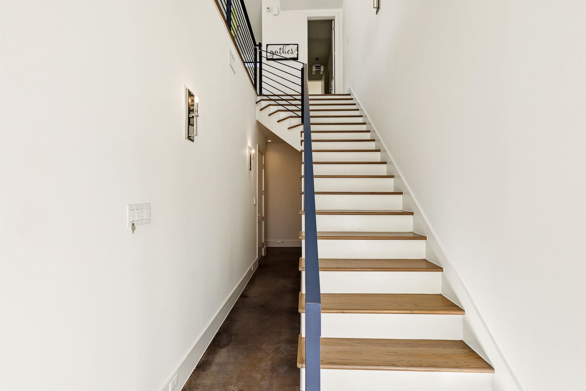 The interior entryway features a straight architectural staircase serving as the focal point, crafted with warm natural-wood treads contrasted against clean white risers. It is framed by a sleek, dark-finished banister and an open horizontal metal railing along the upper landing.
