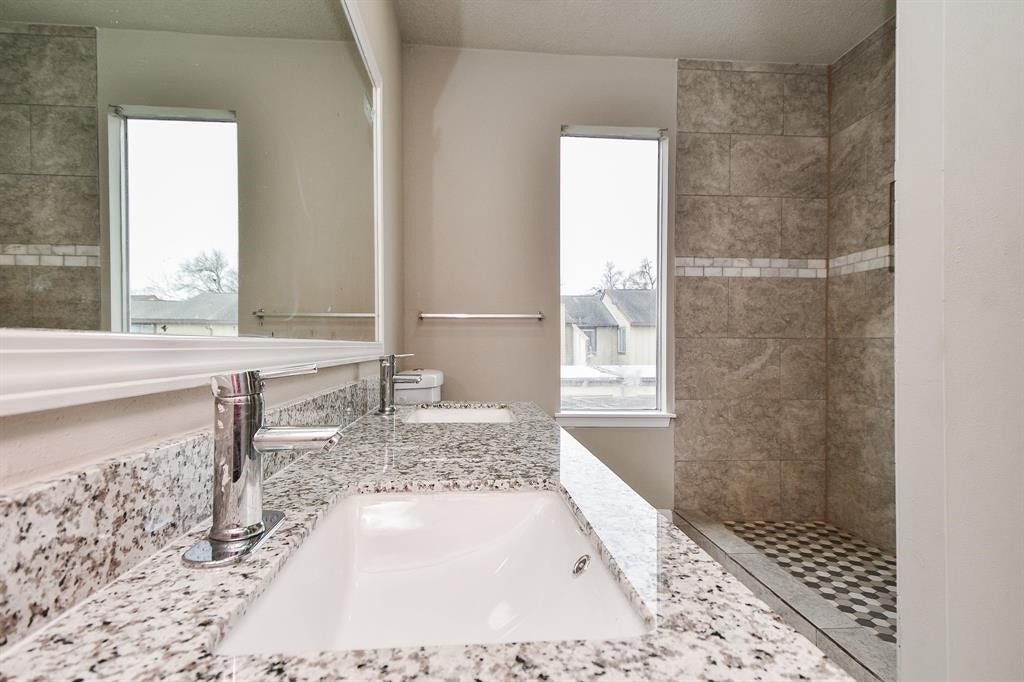 12569 Wellington Park Drive, Houston, TX 77072 Modern bathroom featuring a large granite countertop with dual sinks, sleek fixtures, and a spacious walk-in shower with elegant tilework. Natural light fills the space through two tall windows.